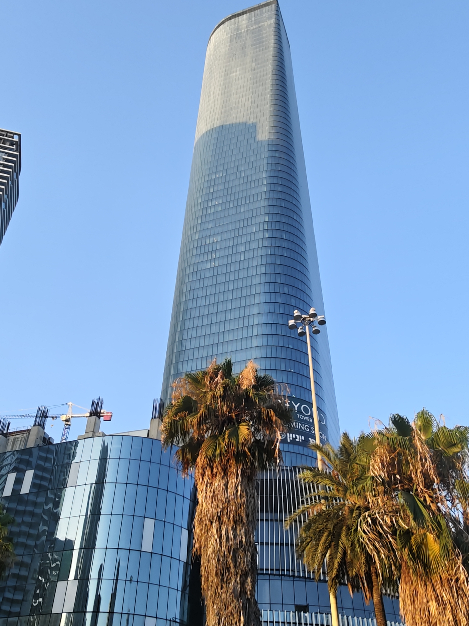 The image shows a view of the OYO Tower under construction in Givatayim, Israel, against a clear blue sky. The tower is a modern, glass-facade skyscraper with a distinctive tapered shape, appearing to be near completion at its upper levels and still under construction at its lower levels, indicated by exposed rebar and construction cranes visible on the left. Palm trees are in the foreground, partially obscuring the lower sections of the buildings. Text on the tower reads "OYO TOWER COMING SOON" in English and Hebrew. A street lamp stands in front of the tower.