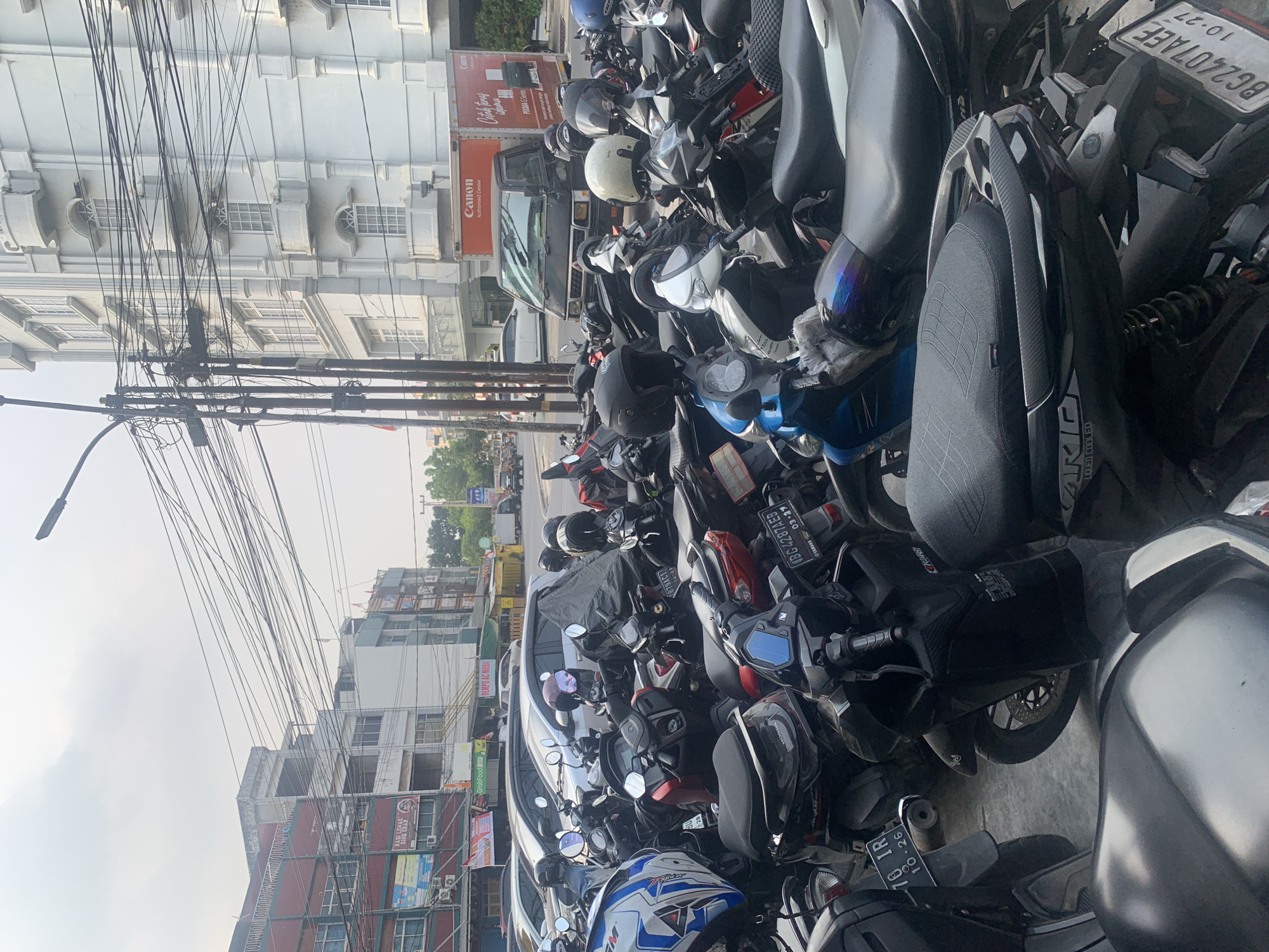 The image depicts a street scene in Palembang, Indonesia, likely near a commercial area. The foreground is dominated by a dense parking lot filled with numerous motorcycles, many of which are covered or have helmets placed on them. Several motorcycles are parked closely together, suggesting a busy area.
In the background, there are buildings lining the street. To the left, multi-story buildings with a red and white facade are visible, with various signs and advertisements displayed. Further back, more buildings extend along the street. To the right, a tall, white building with architectural details like balconies and arched windows stands prominently. A black truck with a "Canon Authorized Dealer" logo on its side is parked in front of this building.
The sky is overcast with a hazy or cloudy appearance. A complex network of electrical wires crisscrosses above the street, suspended from utility poles. The ground appears to be paved. The overall impression is a typical urban street scene in Indonesia, characterized by crowded motorcycle parking, commercial buildings, and overhead utility infrastructure. No people are clearly visible in the image.