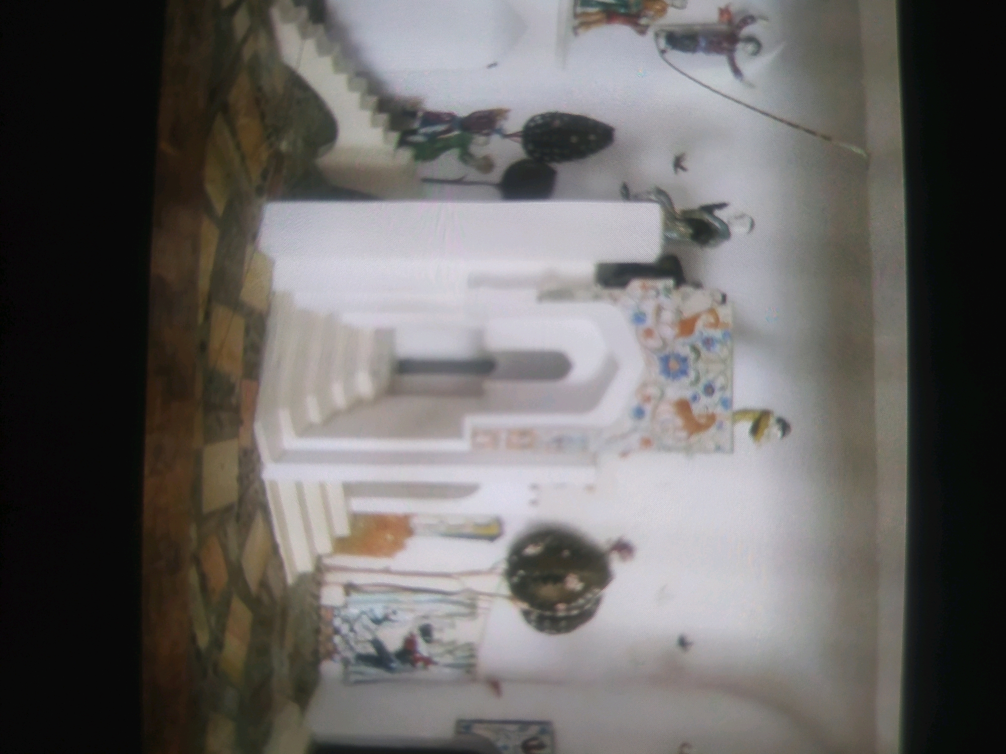 The image shows an indoor scene with a white staircase and decorative elements. The walls are white, and there are some colorful floral decorations on the right side. In the foreground, there is a patterned floor. On the wall, there are several small figures, possibly figurines or toys, in various colors. One prominent object is a large, dark, round decorative item hanging from the ceiling. The scene appears to be a display or an artistic installation. The location is Birlik, Uzbekistan.