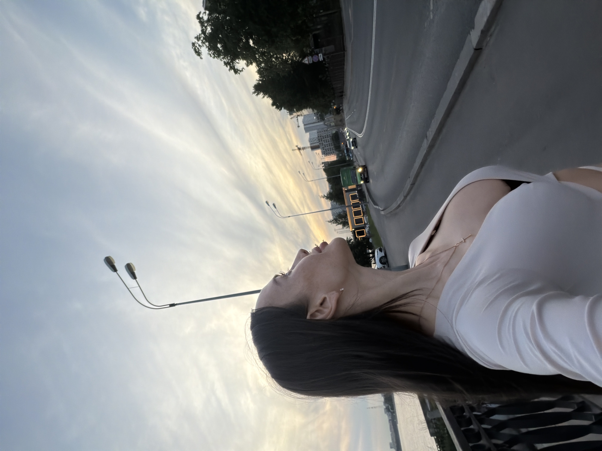 This image features a young adult female in a left-profile view, gazing upwards and to her right, against an urban twilight sky. She has long, dark hair and is wearing a light-colored, long-sleeved top with a v-neckline, revealing a dark undergarment and a gold-colored chain with a small cross pendant. A small, clear stud earring is visible in her right ear.
The scene is located in the city of Saint Petersburg, Russia. To the immediate left of the female, a dark metal railing separates her from a body of water, likely a river or canal, with distant structures visible on the opposite bank. To her right, a multi-lane asphalt road curves into the background, accommodating several vehicles, including a green box truck and a white sedan. Multiple streetlights with twin luminaires are positioned along the road. The roadside includes a pedestrian area, partially lined with trees. In the distance, a collection of multi-story buildings, some under construction with visible cranes, forms the urban skyline. The sky is characterized by elongated, wispy clouds, displaying a gradient of light blue transitioning to soft orange and yellow tones at the horizon, indicative of either sunrise or sunset.