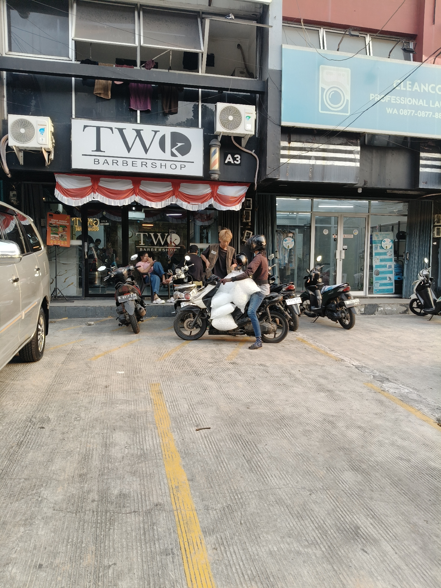 The image shows a street view in front of a barbershop called "TWO BARBERSHOP" and a laundromat called "CLEANCO PROFESSIONAL LAUNDRY" in South Tangerang, Indonesia. Several motorcycles are parked in front of the establishments. A silver minivan is parked on the left side of the image. A person is sitting on a motorcycle, and another person is standing next to it, holding a large white bag. The barbershop has a red and white striped awning. A sign with "A3" is visible on the building. The ground is paved with concrete and has yellow lines indicating parking spaces. The lighting suggests it is daytime, possibly late afternoon.