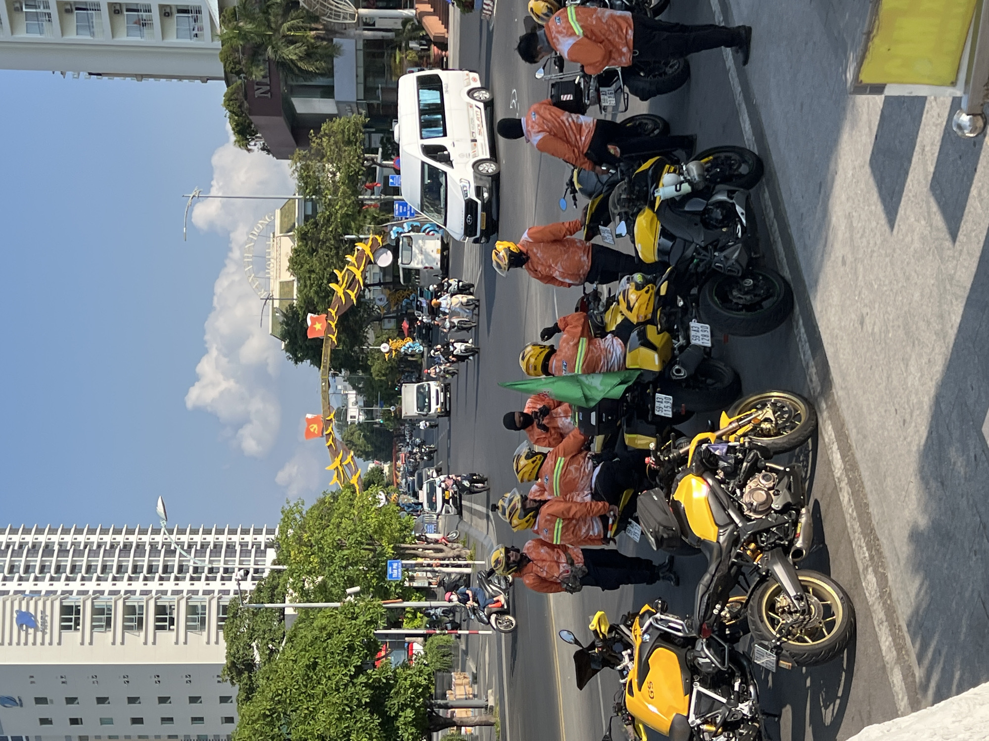 The image shows a street scene in Nha Trang, Vietnam. The perspective is from an elevated position. A row of parked yellow and black BMW motorcycles is arranged along the right side of the frame, with people wearing orange jackets standing nearby. In the background, there is a street with various vehicles, including a white van and other motorbikes. Buildings are visible on both sides of the street, including a tall white building on the left and several other structures further down the road. Vietnamese flags are displayed along the street. The "IXIXIXIX KHANH HOA HOTEL" sign is partially visible in the distance. The sky is blue with some clouds.