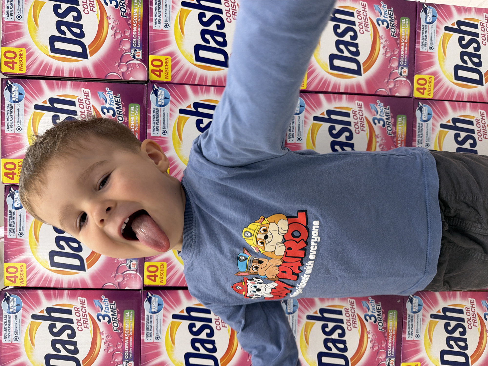 An indoor, rotated image, likely a selfie or close-up, features a young child in Budva, Montenegro. The child, a boy, playfully sticks out his tongue and looks directly at the camera with a joyful, mischievous expression. He wears a light blue long-sleeved shirt adorned with a graphic of "PAW PATROL" characters (Chase, Rubble, Marshall) and the accompanying text "friends with everyone". Parts of his dark gray pants are also visible.
The background consists of a tall, neat stack of "Dash Color Frische" laundry detergent boxes, suggesting a supermarket or retail store aisle. The boxes feature prominent pink, purple, and yellow packaging. Visible text on the boxes includes the product name "Dash Color Frische", "3 fach FORMEL" (3-fold formula), and "COLORWASCHMITTEL Lessive couleur, Detersivo Colore" (color detergent in multiple languages). Each box indicates "40 WÄSCHEN" (40 washes) and highlights environmental claims such as "100% RECYCELBAR" (100% recyclable), "99% PLASTIKFREI" (99% plastic-free), and "Klimaneutral" (climate neutral), often with the hashtag "#VISION PLASTICFREE". The entire scene is presented as if rotated 90 degrees counter-clockwise, with the child appearing sideways.
