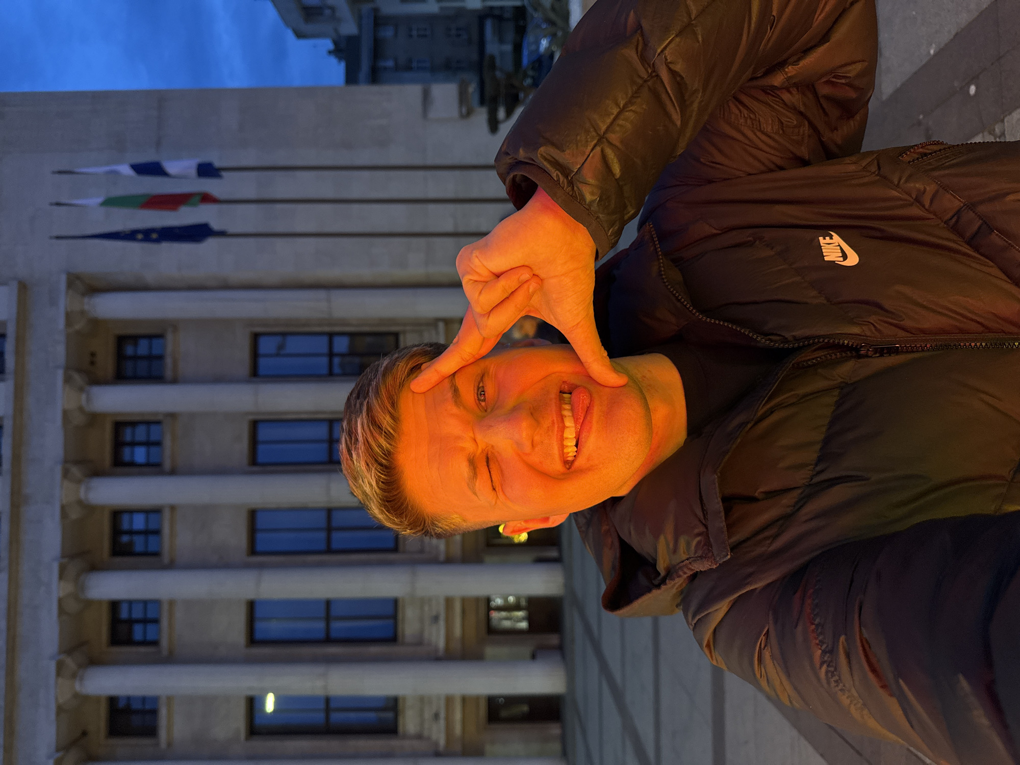 A man in a black Nike puffy jacket stands on a sidewalk in Burgas, Bulgaria, facing the camera while making a triangular shape with his hands around his eyes and sticking out his tongue. Behind him is a large, light-colored building with multiple columns and windows. Three flags—one white and blue, one green and red, and one dark blue with yellow stars—hang from the building. The sky is visible in the background.