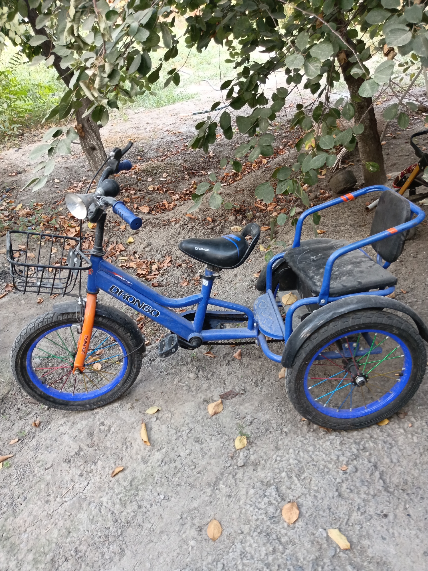 A blue and orange tricycle is parked on a dirt path in an outdoor setting, possibly a park or backyard, in Khodjaobad, Uzbekistan. The tricycle has two seats, one in the front for the rider and a larger one in the back for a passenger. The front wheel is orange and blue, with colorful spokes, and a black basket is attached to the handlebars. The back wheels are also blue with multi-colored spokes. Trees with green and brown leaves are visible in the background, along with fallen leaves scattered on the ground. The overall lighting suggests it is daytime.
