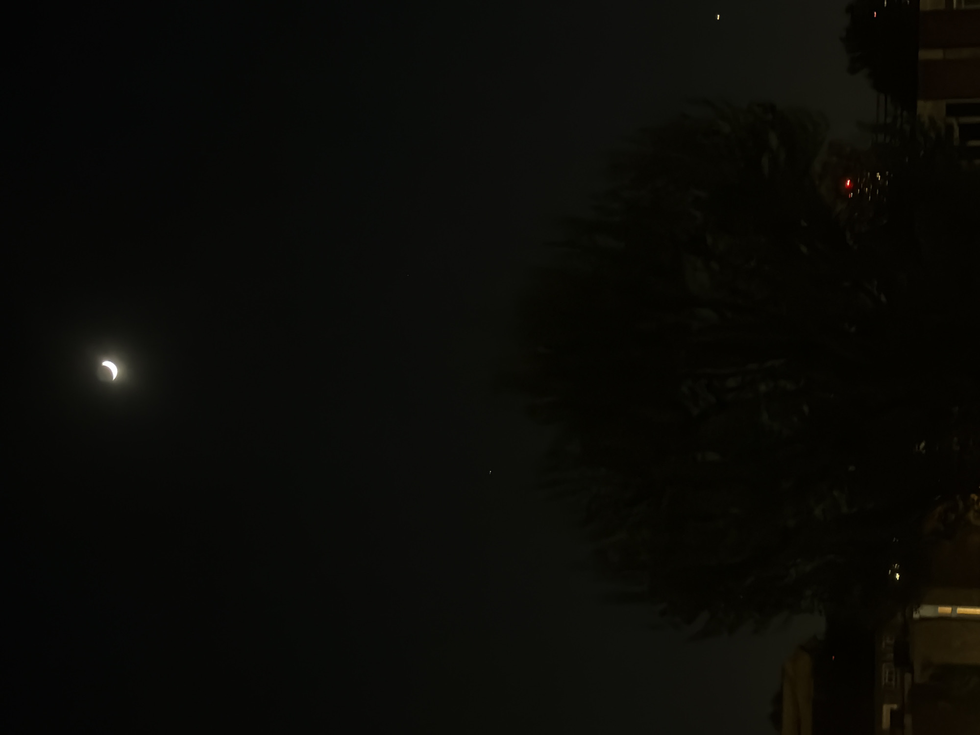 The image shows a night scene with a crescent moon in the upper left. To the right is a dark shape that appears to be a tree. In the upper right, there's a suggestion of a building with some lit windows. No people are visible. The environment appears urban, possibly London, given the description.