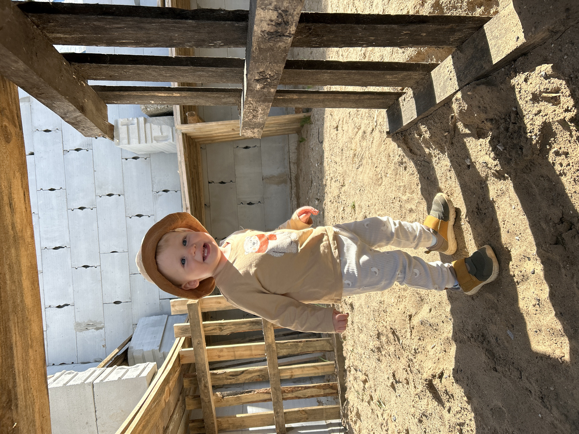 A toddler wearing a light brown cowboy hat, a long-sleeved shirt with a bear image, and striped pants stands outdoors. The child is smiling, with visible teeth, and wears sneakers with green and tan details. The ground is sandy and there is wooden construction material around the child, including what appears to be a staircase structure. In the background, there are light grey building blocks, possibly part of a foundation. The location is likely Chaikovsky, Russia, given the presence of typical construction materials and the style of building. The lighting indicates it's daytime, with strong shadows cast across the scene.