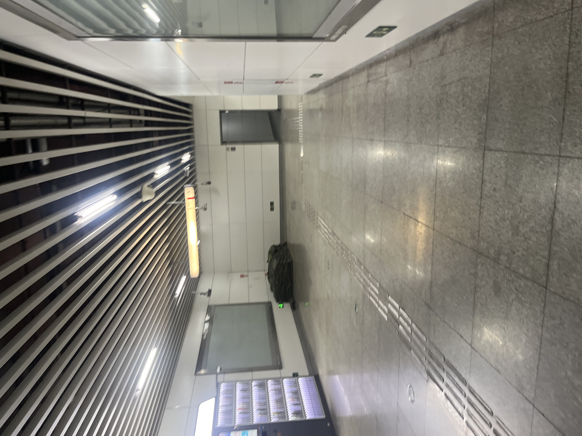 An indoor scene, likely a subway station in Shapingba District, China, depicts a wide, clean hallway with a tiled floor and a modern suspended ceiling system. On the left, a vending machine with illuminated shelves displays products. Adjacent to it, a large, blank digital screen is mounted on the wall. In the center of the hallway, a pile of dark-colored, large sacks is situated. Further down the hallway, a doorway leads to a darker area. The walls are white and panelled, with some signage visible. Tactile paving is present on the floor, indicating a pathway for visually impaired individuals. The lighting appears to be artificial, with recessed lights and a long fluorescent fixture overhead. The overall impression is one of a functional and well-maintained public space. There are no people visible in the frame.
