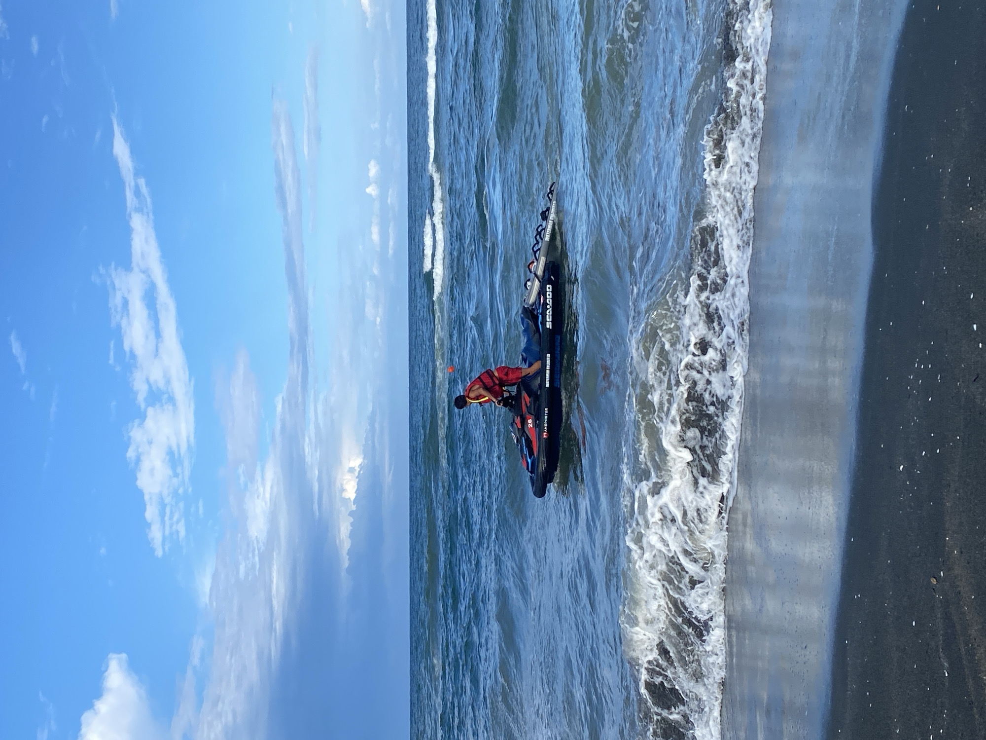 In the city of Ureki, Georgia, on a beach with dark sand, a person wearing a red life vest and red shorts stands on a black and red jet ski in the choppy blue water. The jet ski has "SEA-DOO" written on its side. The sky is blue with scattered white clouds, and waves are crashing on the shore in the foreground.