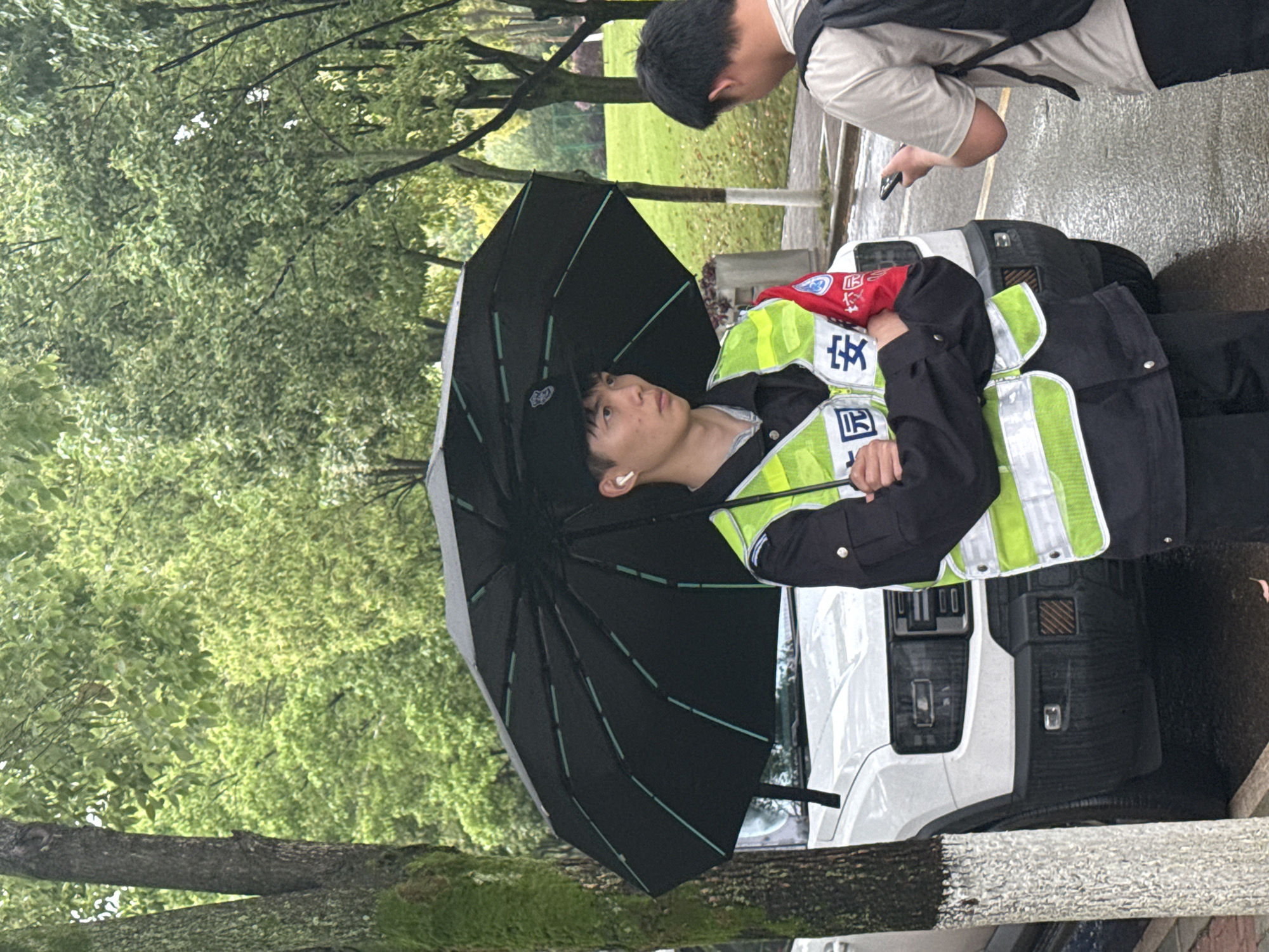 The image shows a person in a security guard uniform, standing next to a white vehicle under an umbrella. The guard is wearing a high-visibility yellow vest with Chinese characters visible on it. Another person, possibly a student, is looking at a phone in the background. The scene appears to be outdoors, with trees and wet pavement visible. The location is likely Jiangjin, China, based on the Chinese characters on the uniform. The light suggests it is daytime, probably overcast due to the umbrella being in use.