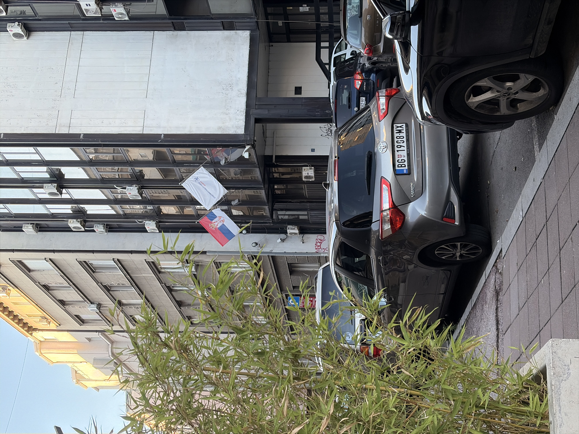 A street view in Belgrade, Serbia, features a modern building with a glass facade and air conditioning units mounted on its exterior. Two flags, one Serbian and one white with an inscription, are displayed. In the foreground, several parked cars are visible, including a silver Toyota Yaris with a license plate reading "BG 1988-MX", a dark blue Suzuki, and a brown car. A tall bamboo plant in a concrete planter is to the left of the frame, partially obscuring the view of the cars and building. The sky is clear and the sun appears to be setting, casting a warm light on the building. The street is paved with cobblestones and asphalt.