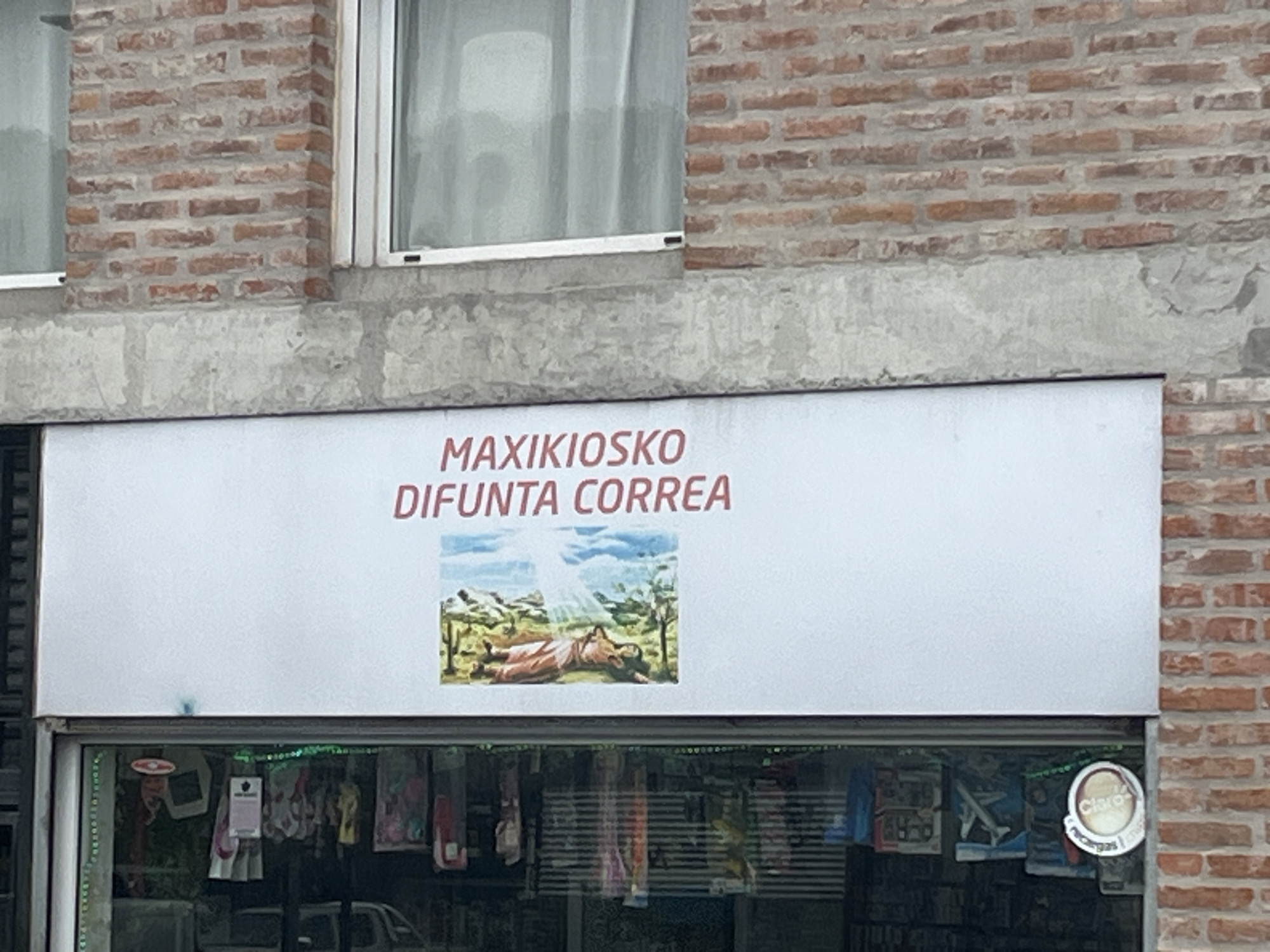 A "Maxikiosko Difunta Correa" convenience store sign is affixed above a window display of merchandise. The sign includes a painting of Difunta Correa, a folk saint. The store is located in a brick building with a concrete overhang. A window with sheer curtains is visible above the sign. The store's window display is decorated with green garland. A "Claro" cellular recharge sign is affixed to the window. The location is Buenos Aires, Argentina.