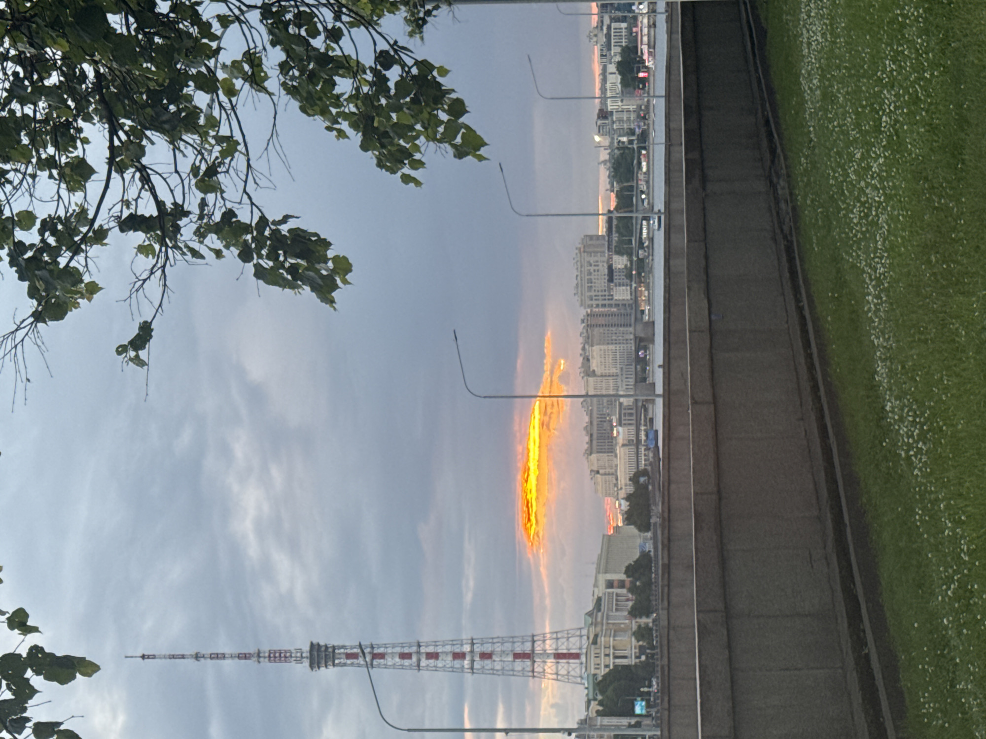 The image depicts a view of the city of Saint Petersburg, Russia, across a river, likely during sunset. In the foreground, there is a concrete embankment and a grassy area with small white flowers. A tall, red and white striped radio tower stands prominently on the left side of the frame, with a cluster of buildings and a bridge visible behind it and to the right. The sky is overcast with dramatic, brightly colored clouds, suggesting the sun is low on the horizon. Several streetlights are visible along the embankment. Trees with green leaves frame the upper right portion of the image. The overall impression is of an urban landscape at dusk.