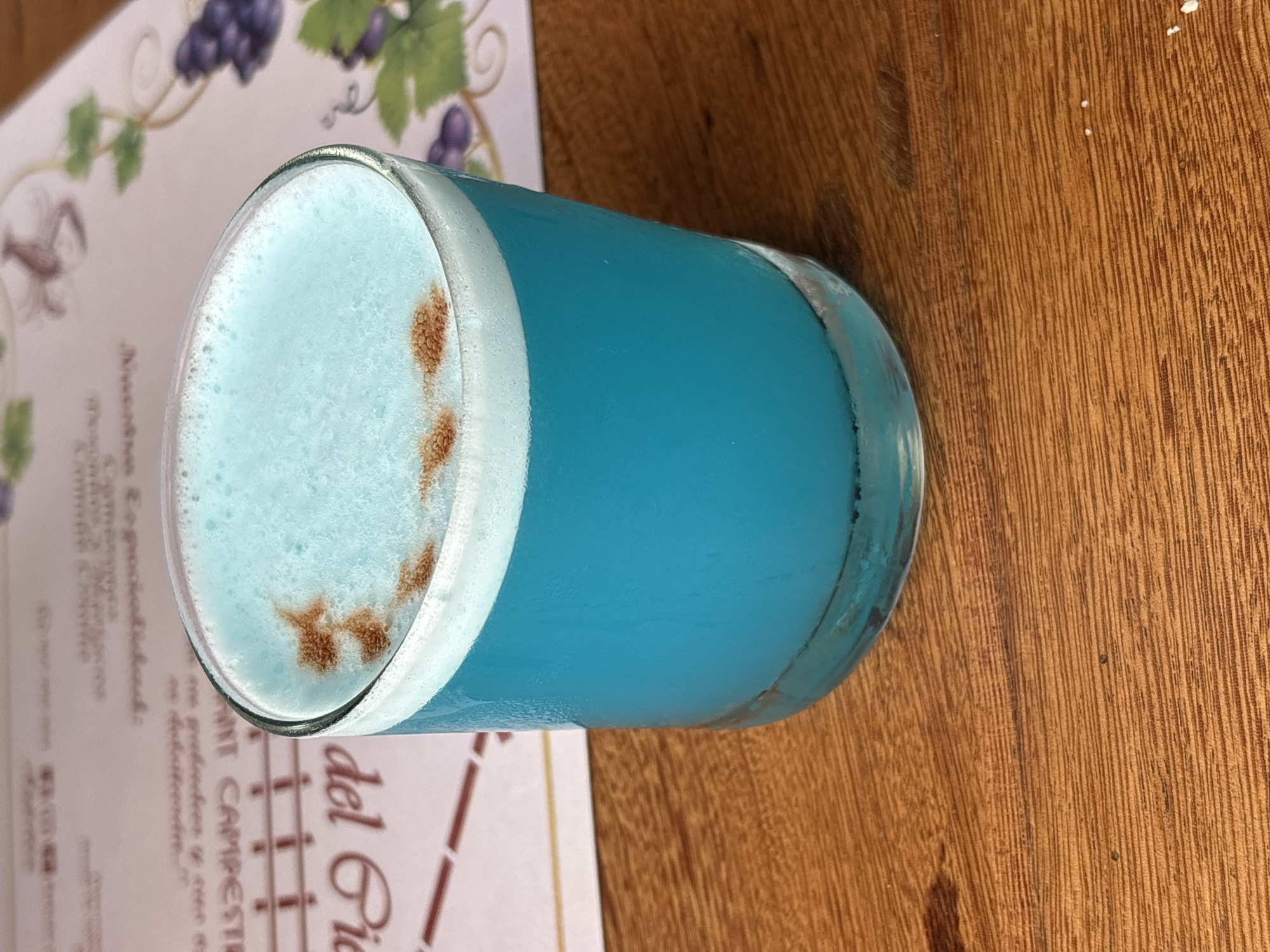 A close-up, eye-level, medium shot captures a tall, clear glass filled with a bright blue beverage, topped with a frothy white foam. The foam is sprinkled with a light dusting of cinnamon in a splotchy pattern. The glass is positioned on a wooden table with a prominent wood grain. In the background, a pinkish-white menu is visible, featuring decorative illustrations of grapevines and grapes on the right side, and what appears to be text and vertical lines on the left. A small portion of the menu reads "del Rio" and "del CAMPESINO". The overall setting suggests a restaurant or cafe in San Vicente de Azpitia, Peru, a region known for its agricultural products and vineyards. The lighting is natural, with a slight shadow cast on the wooden surface to the left of the glass. There are no people present in the frame.