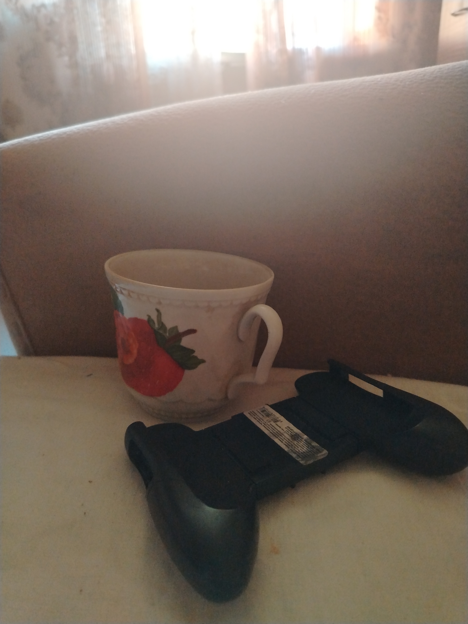 An indoor, eye-level, close-up shot shows a black video game controller resting on a white surface next to a white teacup with a strawberry design. The controller is oriented horizontally, with its handles facing left and right. The teacup is positioned to the left of the controller and is slightly taller than it. The background is blurred, showing a muted brown couch and a window with translucent curtains. The lighting suggests a bright, possibly sunny day, with light filtering through the curtains. The image appears to be taken in a residential setting, likely a living room or lounge area. There are no people or animals visible in the frame. The scene is static and appears to be a still life arrangement. The specific location in Kirov, Russia, is not discernible from the visual information provided.