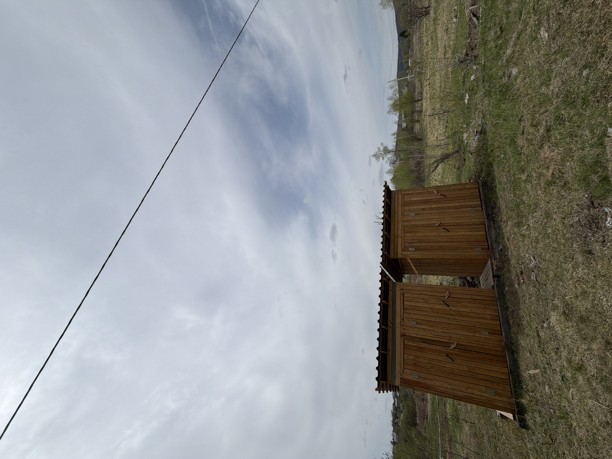 The image shows two wooden outhouses with corrugated metal roofs located in a grassy area, possibly in Теплоозёрск, Russia. A power line is visible in the sky. The area appears to be rural or on the edge of town. The grass is dry in some spots, and the sky is cloudy.