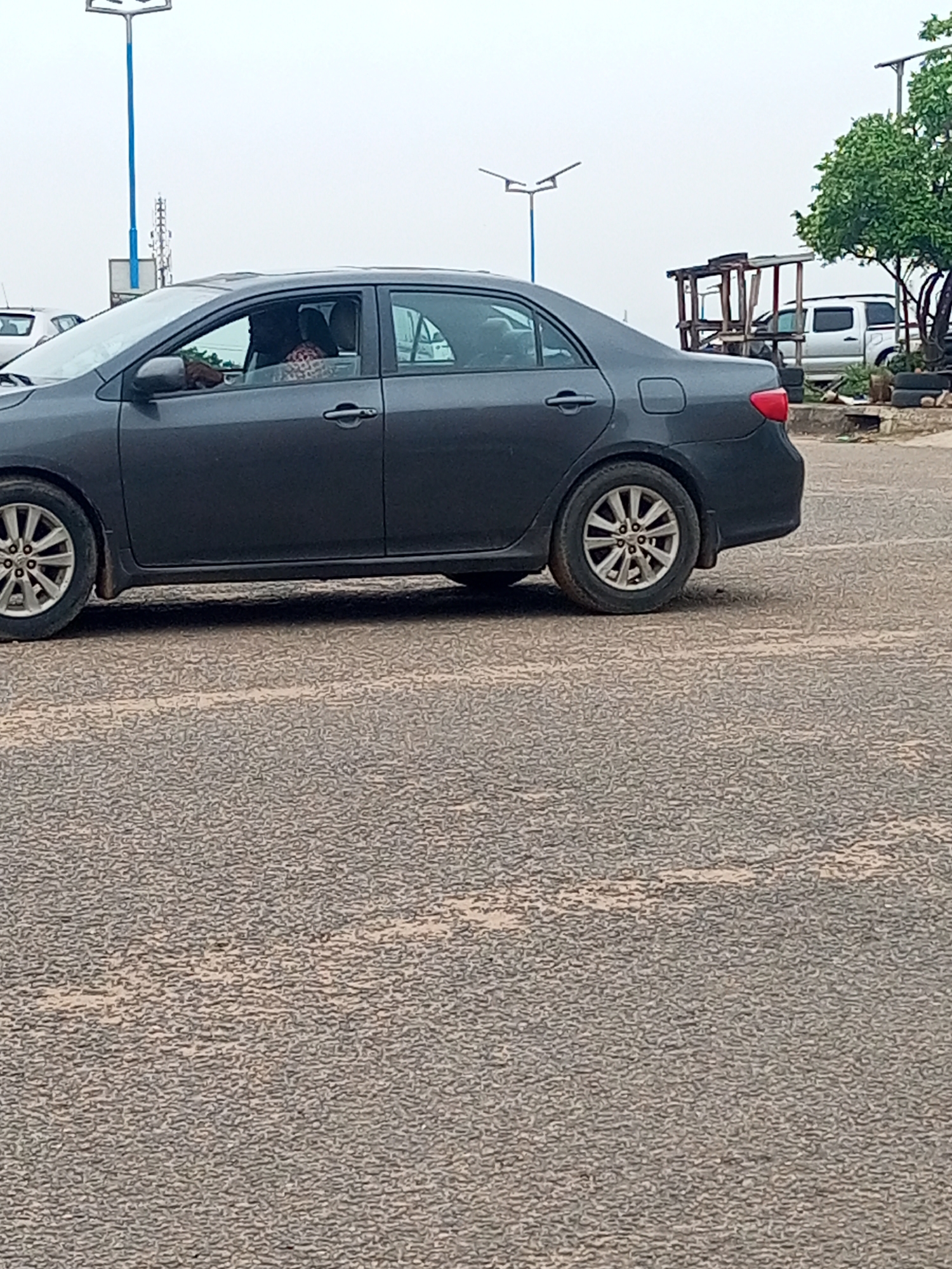A gray sedan is parked on a coarse gray asphalt road in what appears to be Oshogbo, Nigeria, as suggested by the urban setting and the style of streetlights and roadside structures in the background. Inside the car, a person is visible in the driver's seat wearing patterned clothing. Behind the car, a white pickup truck can be seen parked near a simple wooden structure. A small tree is next to the wooden structure. Tall streetlights are also visible in the background.