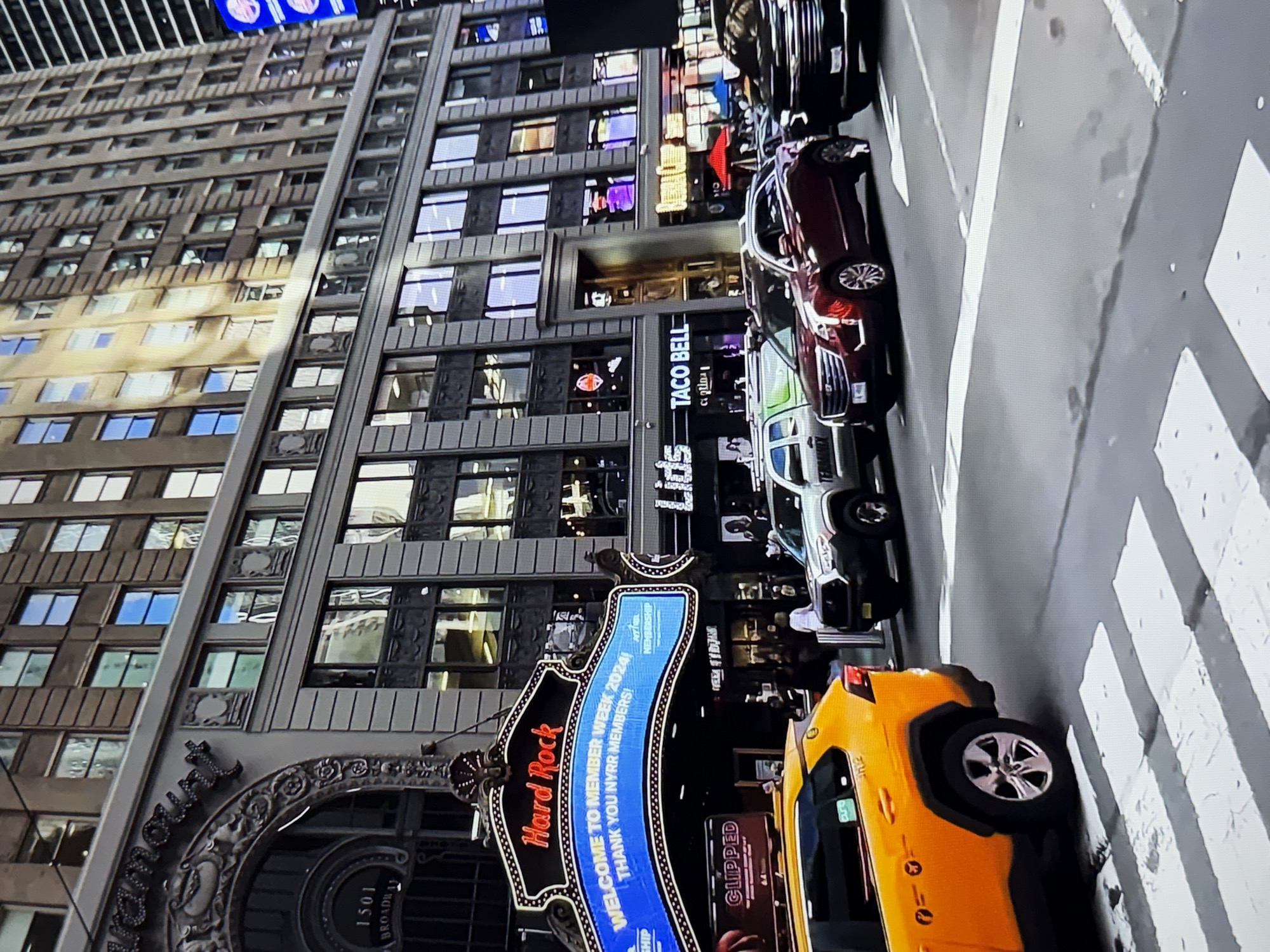 This is a medium-shot, eye-level, full-color, slightly blurry photograph of a street scene in New York City, USA, not Burgas, Bulgaria. The Hard Rock Cafe marquee is prominently displayed, featuring a blue LED screen that reads "WELCOME TO MEMBER WEEK 2024! THANK YOU NYRR MEMBERS!". The building behind the marquee is a multi-story structure with numerous windows. In the foreground, a yellow taxi cab is visible, and several cars are driving on the street, including a red Cadillac SUV and a black SUV. The scene appears to be set during daytime.