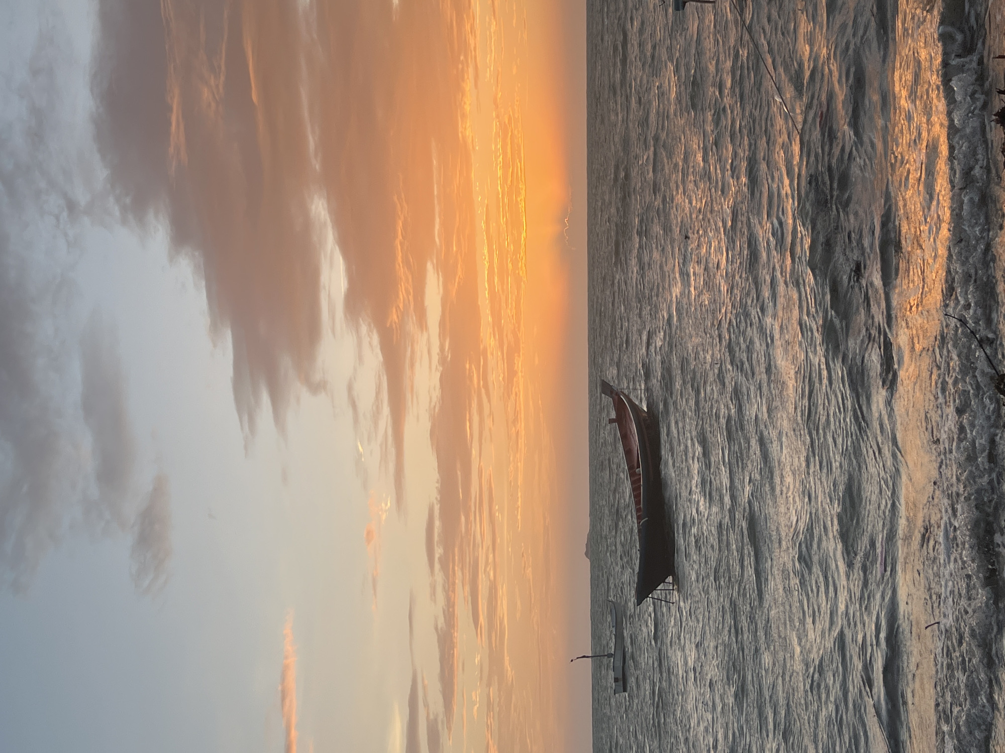 This photograph depicts a serene seascape at sunset in the Ko Pha-ngan District, Thailand. The sky is painted with vibrant hues of orange and yellow, transitioning into softer pinks and blues as it rises. Wispy clouds are scattered across the horizon, catching the last rays of sunlight.
The ocean is calm, with gentle waves lapping at the shore. The water reflects the warm colors of the sunset, creating a shimmering surface. Two boats are visible on the water. In the foreground, a dark-hulled boat with red trim rests peacefully. Further in the distance, a smaller, lighter-colored boat with a tall mast bobs on the waves.
A hint of land is visible on the far horizon, suggesting the presence of distant islands or the mainland. The overall atmosphere is one of tranquility and natural beauty, characteristic of a tropical sunset.
