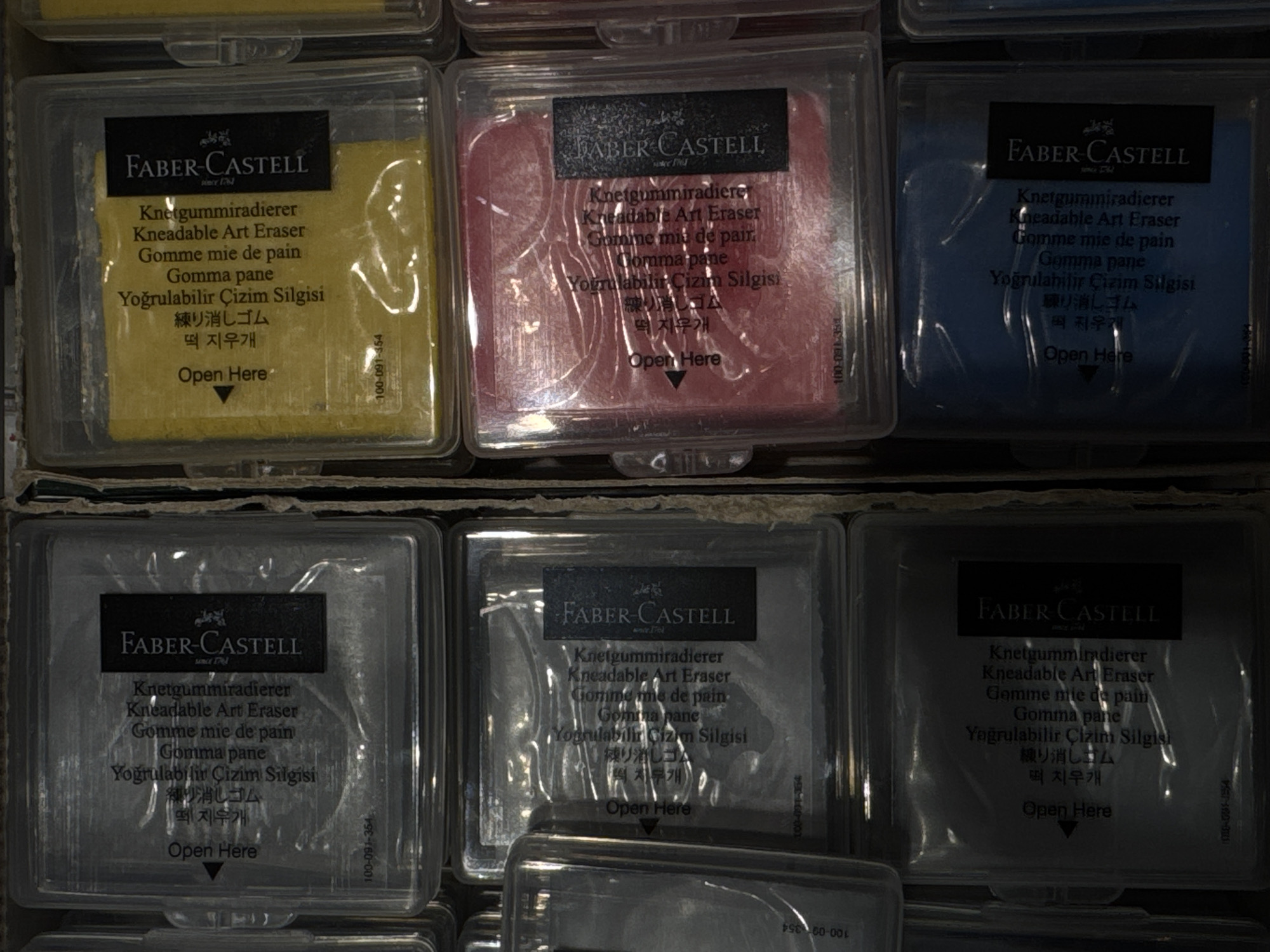 This image displays a collection of Faber-Castell kneadable art erasers arranged in a grid of six square, transparent plastic containers. The erasers themselves are visible within their packaging and appear in various colors: yellow, pink, blue, and grey. The containers are stacked within a larger cardboard box, suggesting a retail or storage environment. Each container has a label with the "FABER-CASTELL" logo and product information in multiple languages, including English, German, French, Italian, Turkish, Japanese, and Korean. Text visible on the labels includes "Knetgummiradierer," "Kneadable Art Eraser," "Gomme mie de pain," "Gomma pane," "Yoğrulabilir Çizim Silgisi," "練り消しゴム," and "떡 지우개." Some containers also have numerical codes printed on them, such as "100-091-354." The lighting is somewhat dim, casting shadows and creating a mood of focused presentation of the art supplies. The scene is set indoors, likely in a store or warehouse. No people are visible in the image.
