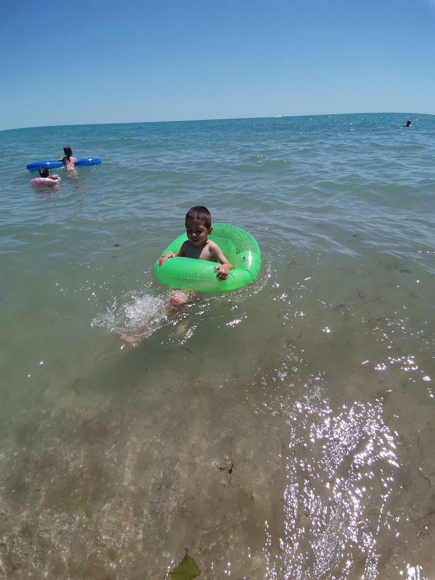 A child is in the water wearing a green inflatable ring. The child is looking to their right and smiling. There are other people in the water further away, one on a blue inflatable and another on a pink inflatable. The water is clear and the bottom is visible in some areas. The scene is a beach on a sunny day with a clear blue sky. The location is Kranevo, Bulgaria. There is no visible text in the image.