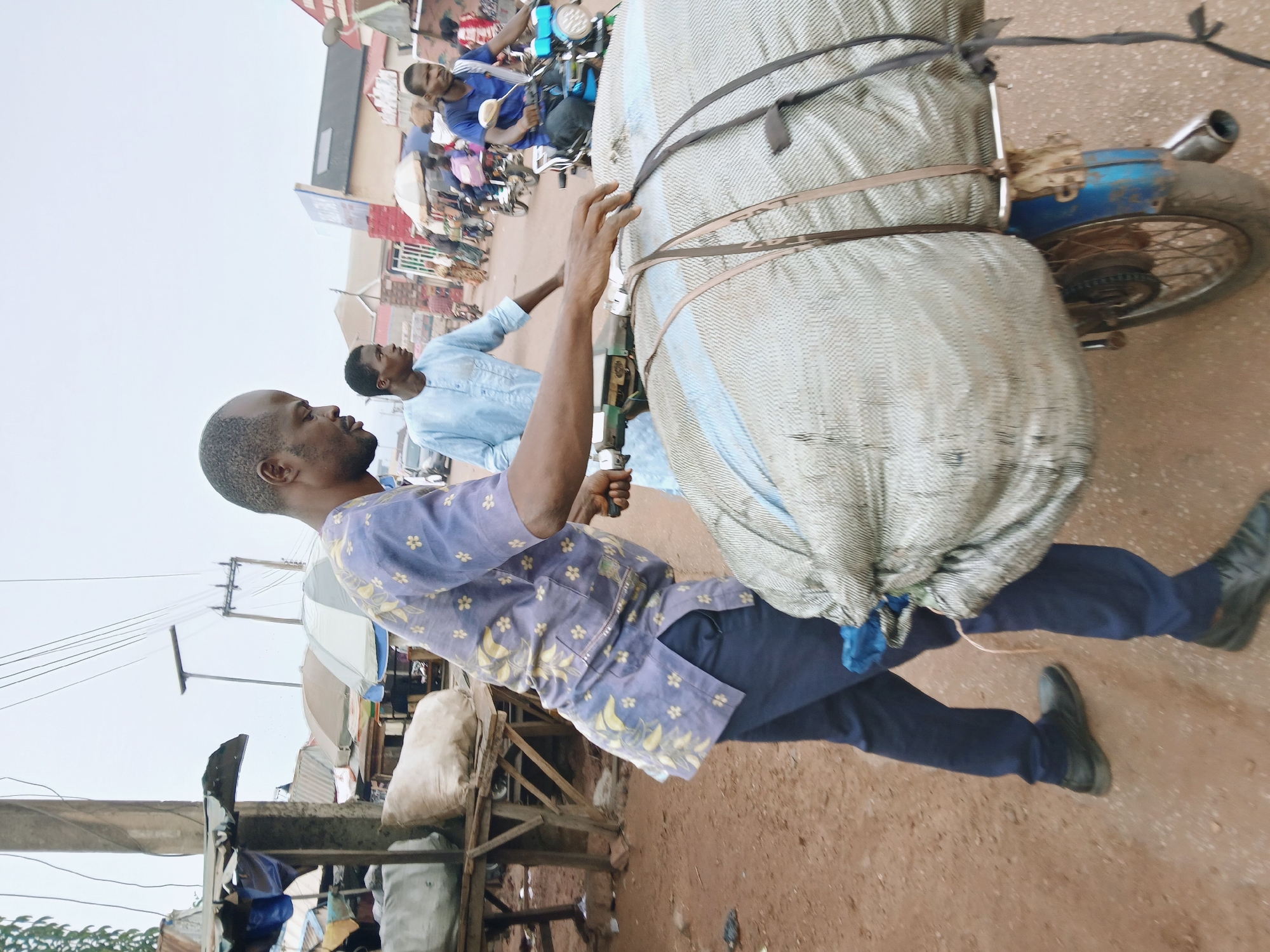 In a street scene in Ankpa, Nigeria, a man wearing a patterned blue shirt and dark pants walks along a dirt road. He is pulling a large, fabric-covered load secured to a blue motorcycle. The man is facing to the left. Another man, wearing a light blue shirt, is walking behind him. In the background, there is a crowd of people near a building. To the left of the walking man, there are makeshift stalls and goods displayed. Overhead power lines are visible.