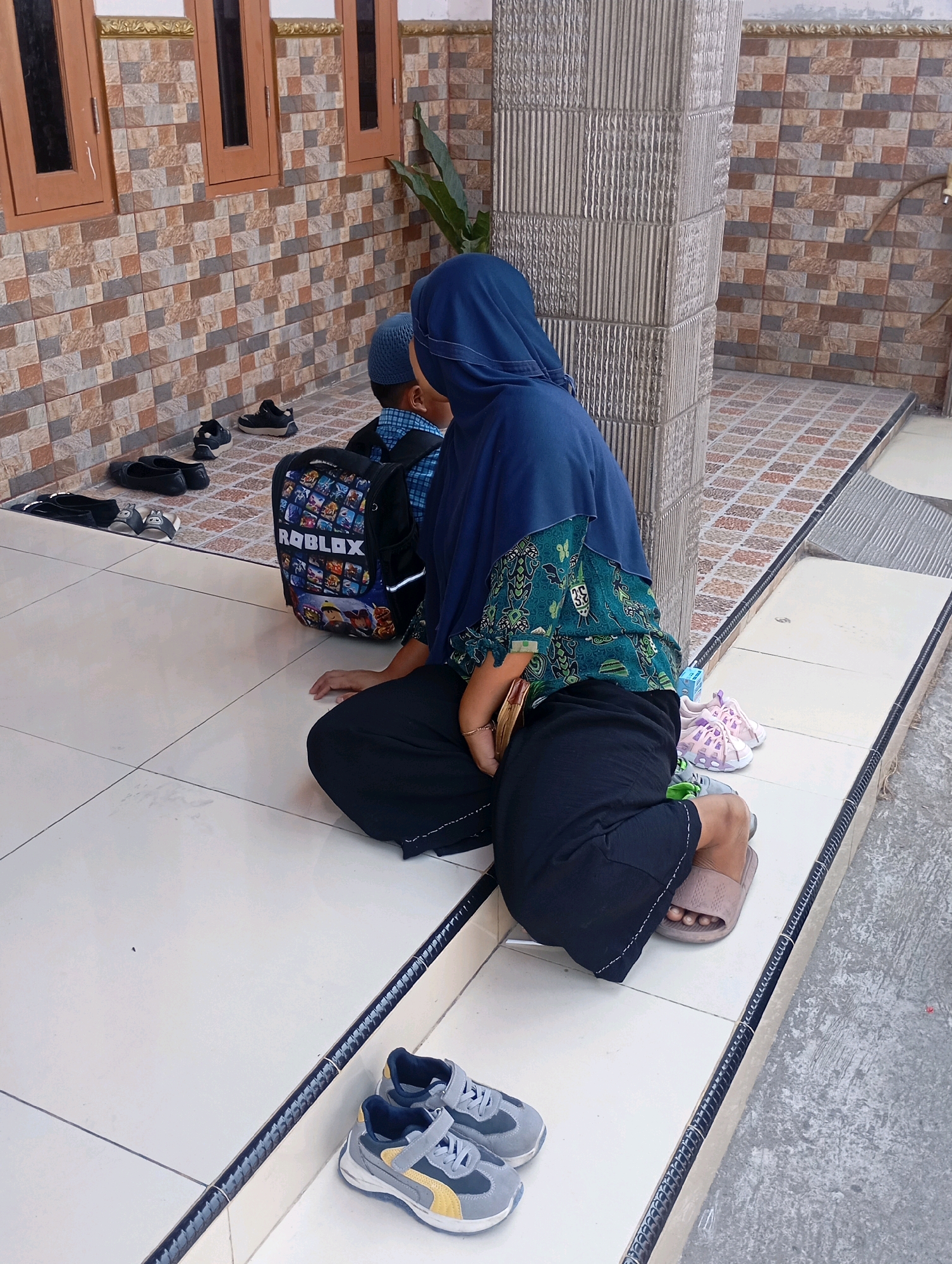 In Babelan, Indonesia, a woman wearing a blue hijab and a patterned green blouse sits on a white tiled floor next to a pillar. She is dressed in black pants and brown slippers. To her right are a pair of pink children's sandals and a small blue box. In front of her sits a child wearing a blue cap and a backpack with "ROBLOX" printed on it. Scattered on the tiled floor are several pairs of shoes, including black slip-on shoes, black sneakers, and gray and yellow children's sneakers. The background features a wall with brown-framed windows and a tiled exterior.