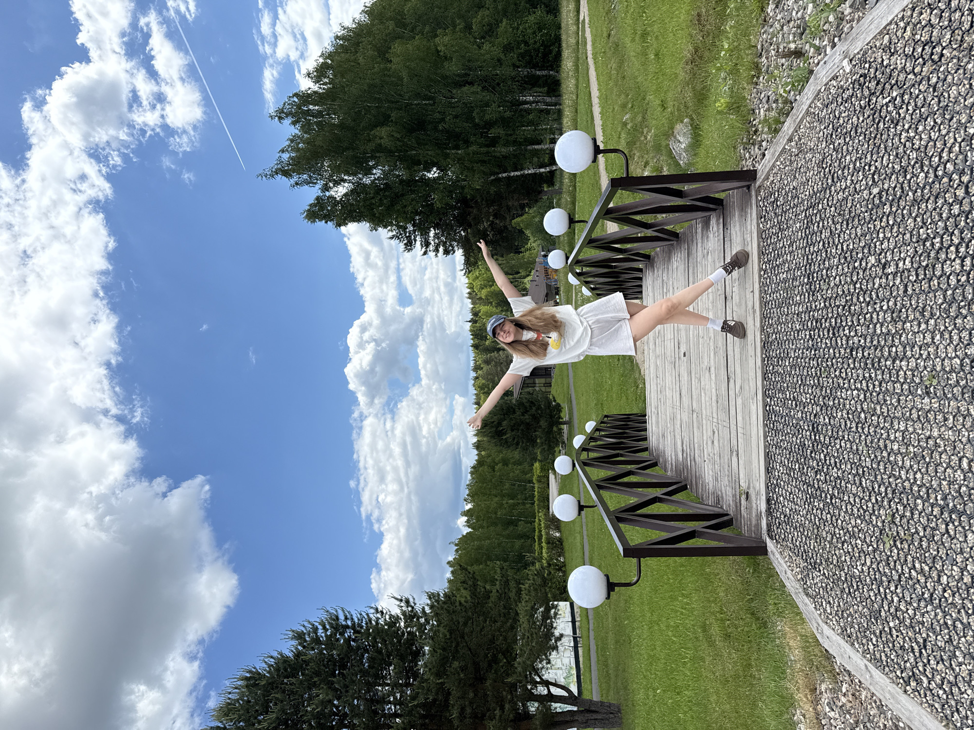 The image displays a young woman posing joyfully on a wooden bridge or walkway in an outdoor setting, likely a park or recreational area, under a bright blue sky. She stands centrally on the walkway with her arms outstretched and legs crossed, smiling directly at the camera. She has long, light brown hair and is wearing a light-colored t-shirt with a small graphic, light-colored shorts, white socks, dark shoes, and a blue baseball cap.
The wooden walkway features dark, X-patterned railings along its side, on which several spherical white light fixtures are mounted. Adjacent to the walkway is a textured path made of dark hexagonal grids filled with light gravel, bordered by green grass. In the background, a dense line of lush green trees, including both deciduous and coniferous varieties, stretches across the horizon.
The sky is a clear, vibrant blue, dotted with scattered white, fluffy clouds. A prominent white contrail from an airplane is visible high in the sky, indicating fair weather and daytime. The overall scene suggests a pleasant, sunny day, with the woman expressing a sense of freedom and enjoyment in a natural environment, possibly in Dobrograd, Russia. There is no visible text in the image.