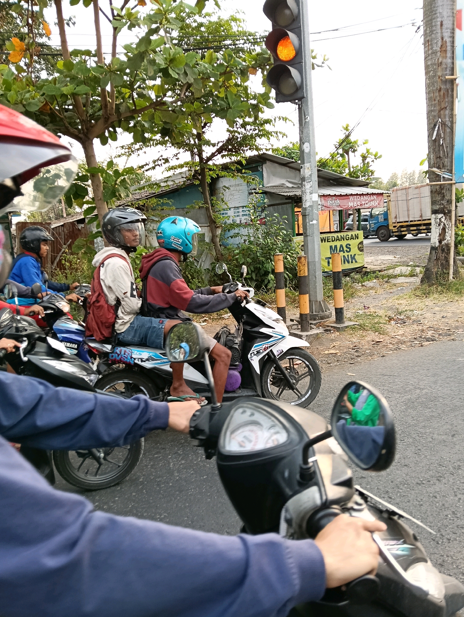 A group of motorcyclists are stopped at a traffic light in Jaten, Indonesia. The traffic light is orange. The motorcyclists are wearing helmets and casual clothing. Some of the motorcycles have passengers. In the background, there are buildings, trees, and a truck. The scene appears to be a typical street in a suburban or rural area of Indonesia.
