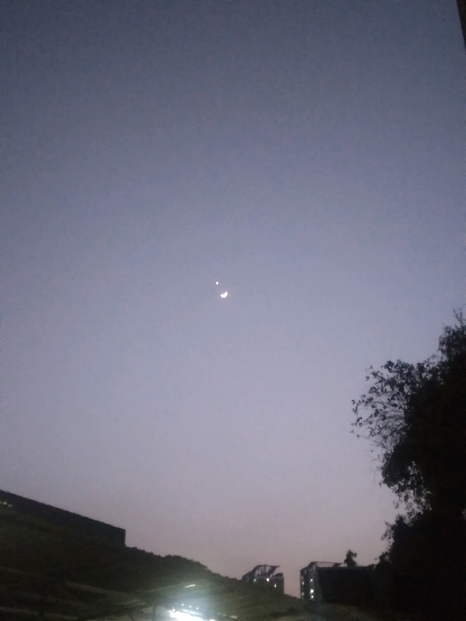 The image depicts a dusky sky over a city landscape, likely South Jakarta, Indonesia. Two celestial bodies are visible in the upper center of the frame; one is a crescent moon, and the other appears to be a planet, possibly Venus, due to its bright, star-like appearance and proximity to the moon. In the lower portion of the image, dark silhouettes of buildings and trees are visible against the fading light. Some lights are visible in the distance, suggesting urban activity. The overall atmosphere is one of twilight.