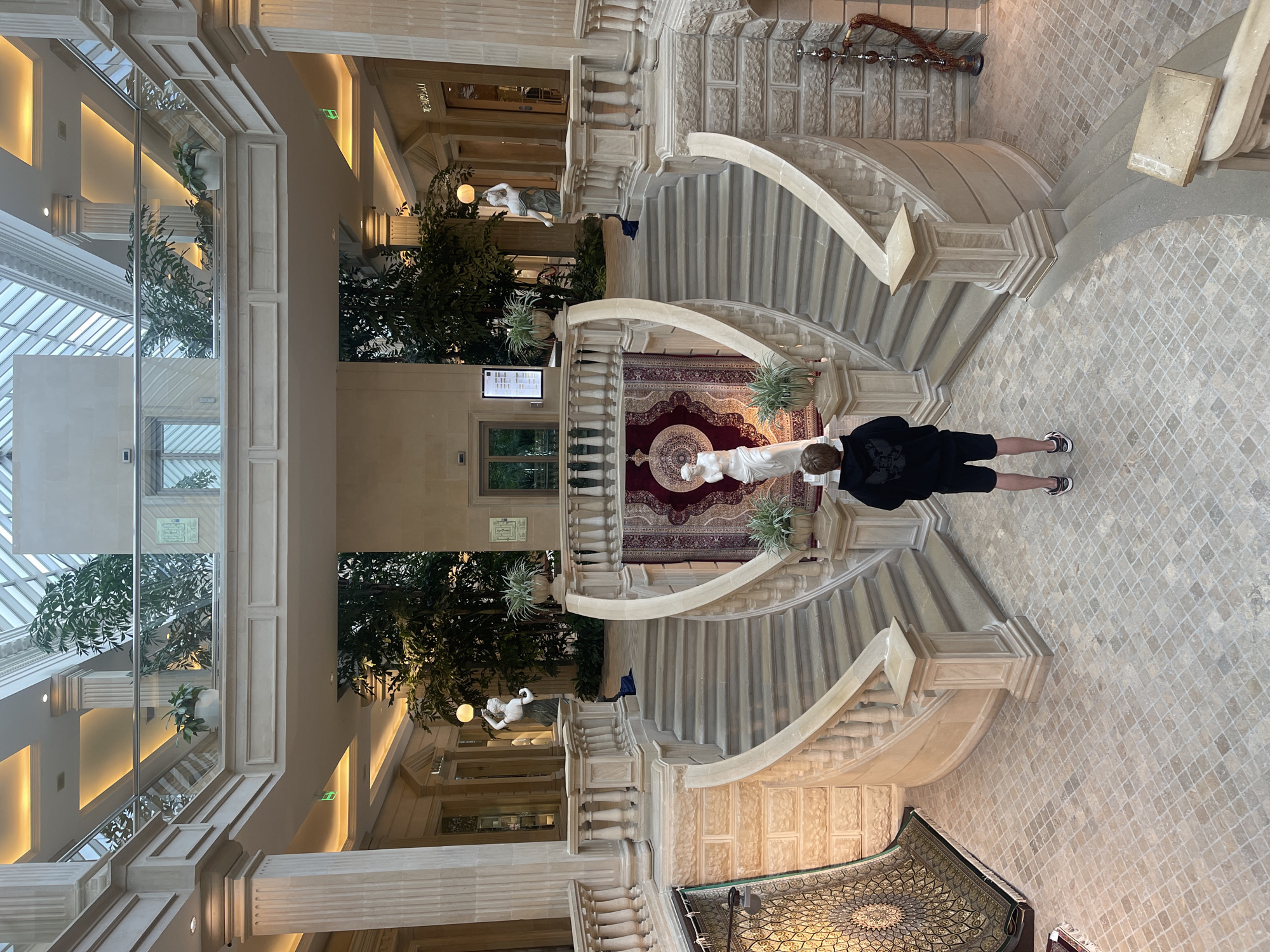 The image shows a grand interior staircase with ornate balustrades and stone steps. A person wearing a black hoodie and shorts stands on the tiled floor, facing away from the camera, near the base of the staircase. The staircase is adorned with a large red rug featuring intricate patterns and a circular medallion. Two white statues are visible along the staircase, one reclining and another partially visible at the bottom. Lush green plants are strategically placed throughout the atrium, adding a touch of nature to the classical architecture. The ceiling features a glass-paneled skylight, allowing natural light to illuminate the space, suggesting daytime. The architecture, with its stone columns and detailed carvings, points to a formal or historical setting, possibly a hotel lobby, museum, or grand residence. There are no visible signs of extreme weather conditions. The overall impression is one of elegance and opulence.