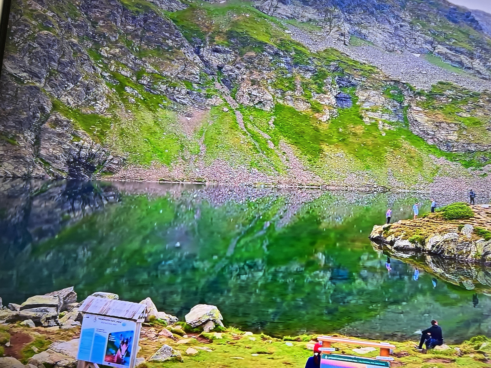 A lake scene with a backdrop of a mountain and a shoreline with people, benches, and signage. The mountain is covered in rocks, and green vegetation. The lake reflects the mountain's scenery. Some people are standing on the rocky shoreline, and one person is seated near a bench. There's a wooden sign with information on it. The location appears to be in a natural, mountainous area. The city and state are unknown.