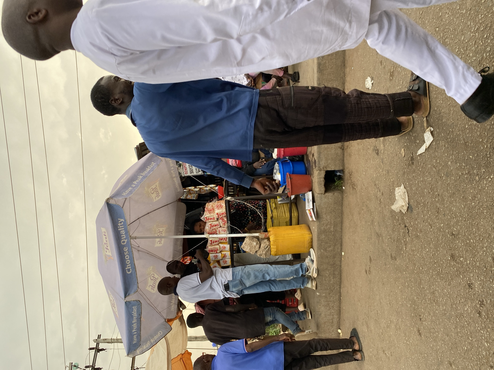 The image depicts a street scene, likely in Bukuru, Nigeria, based on visual cues. A small open-air shop, sheltered by a blue "Peak" branded umbrella, is the focal point. Several people stand in a loose queue in front of the shop, presumably waiting to make purchases. The shop sells an array of packaged goods, likely food items, displayed on shelves. Colored plastic containers are stacked near the shop. The people are dressed in casual clothing. The foreground is occupied by people walking past the shop. The street surface is concrete. Overhead electrical wires are visible.