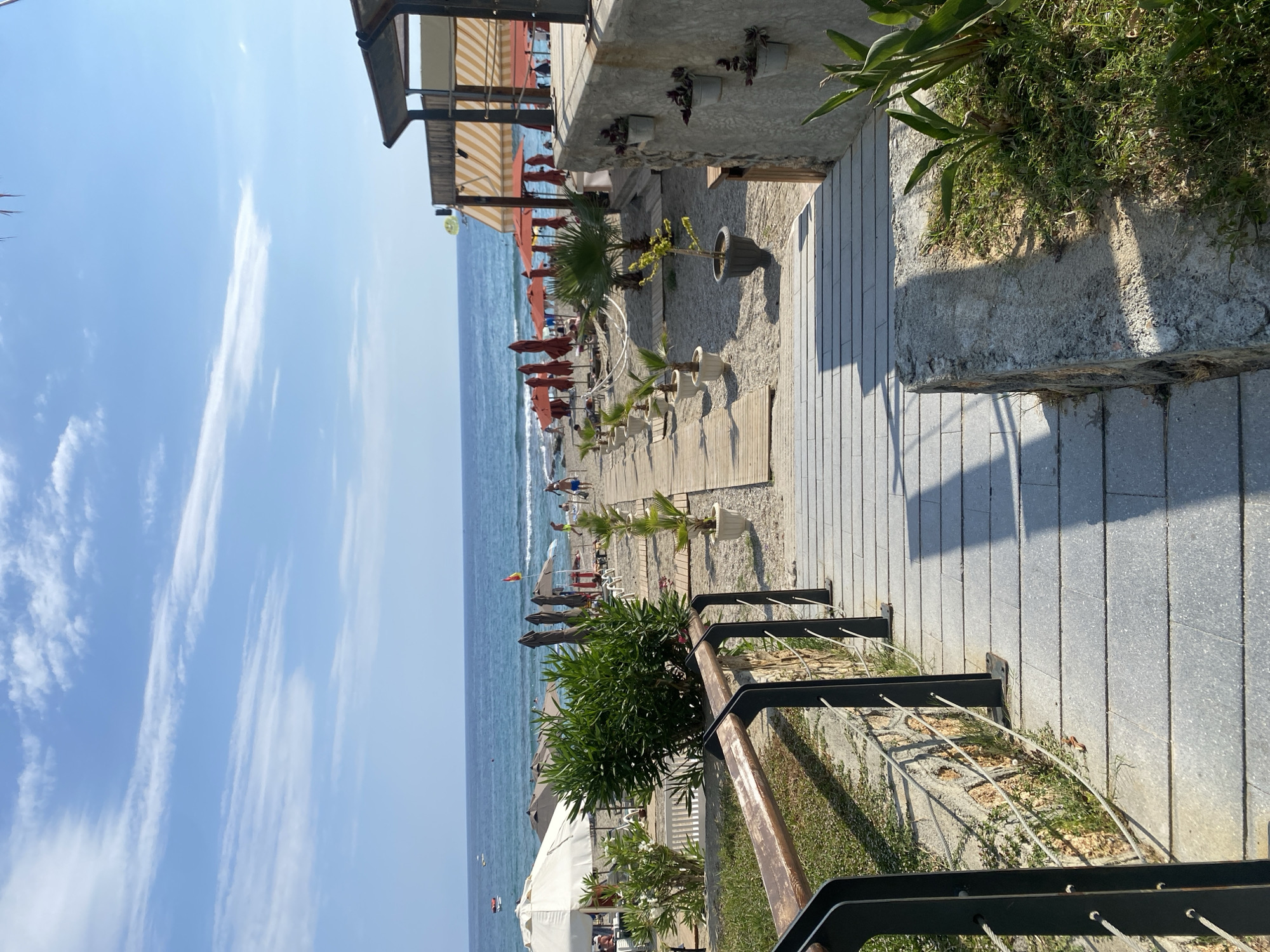 A wooden boardwalk leads down to a sandy beach in Alanya, Turkey, under a bright blue sky with scattered white clouds. The boardwalk is flanked by a railing and a stone wall with small potted plants. On the beach, lounge chairs and red umbrellas are set up. People are visible in the distance, enjoying the water and the sand. The sea is calm and blue, with small waves breaking near the shore. A parasail is visible in the sky. The overall atmosphere is one of a relaxed summer day at a resort.