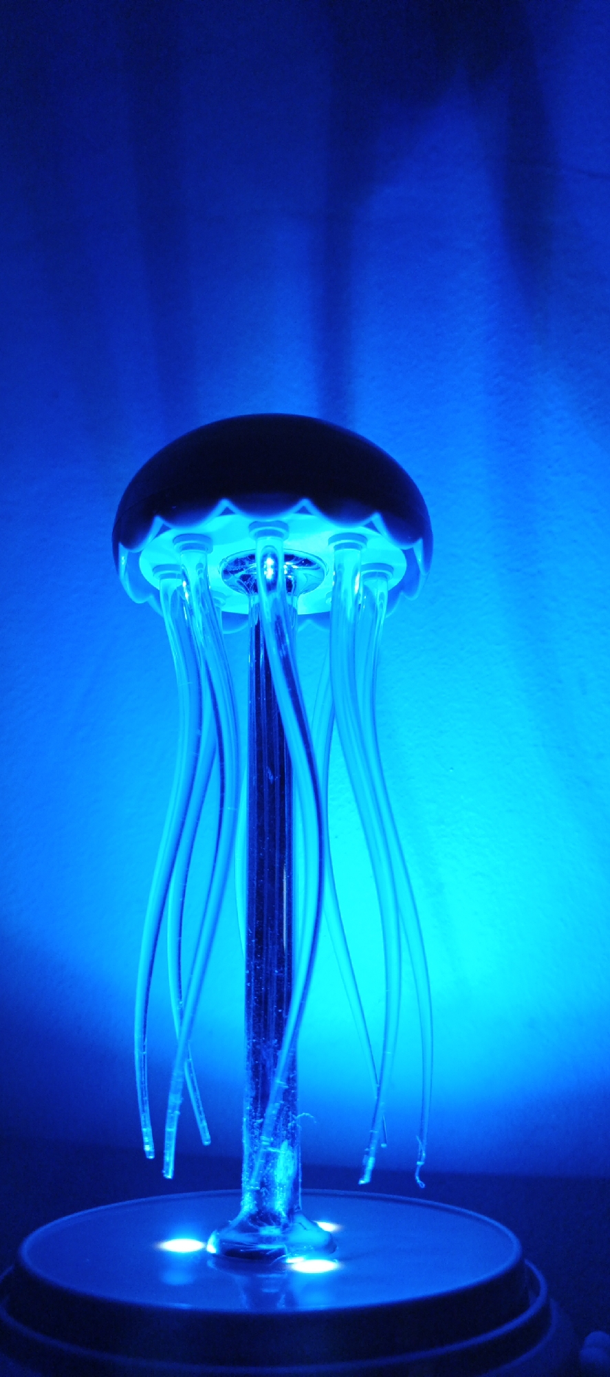 A low-angle, close-up shot features a jellyfish lamp with a dark-colored, rounded top and several clear, glowing blue tentacles hanging down. The lamp sits on a circular base with illuminated white lights. The background is a gradient of blue, with a slightly textured wall visible behind the lamp. The image was not necessarily taken in Chaikovsky, Russia, there is nothing specific in this image that indicates that it was.