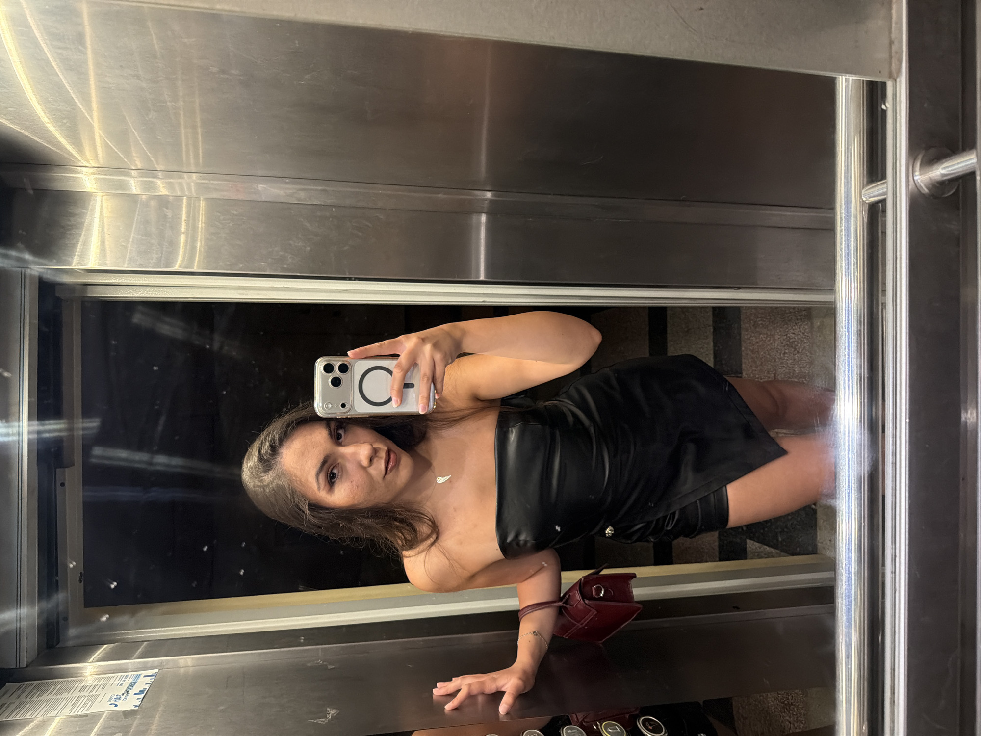 A young woman is taking a mirror selfie in an elevator. She is wearing a black strapless leather-like dress with a wrap-style skirt. She has long dark hair and is wearing makeup, including lipstick. A small red handbag is hanging from her left arm. The elevator has stainless steel walls and doors. The floor appears to be tiled. A sticker with text is visible on the elevator wall. The image was taken in the city of Varna, Bulgaria.