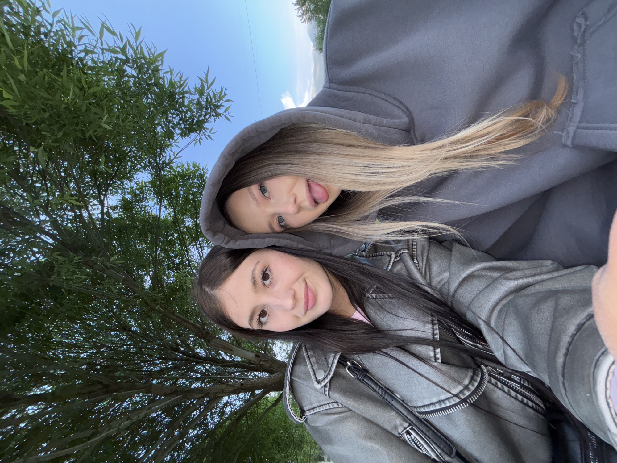 Two young women are posing for a selfie outdoors. One woman is wearing a grey hoodie and has blonde hair; she is sticking her tongue out. The other woman is wearing a grey jacket and has dark hair. Trees and a blue sky are visible in the background. The location is possibly Chaikovsky, Russia.