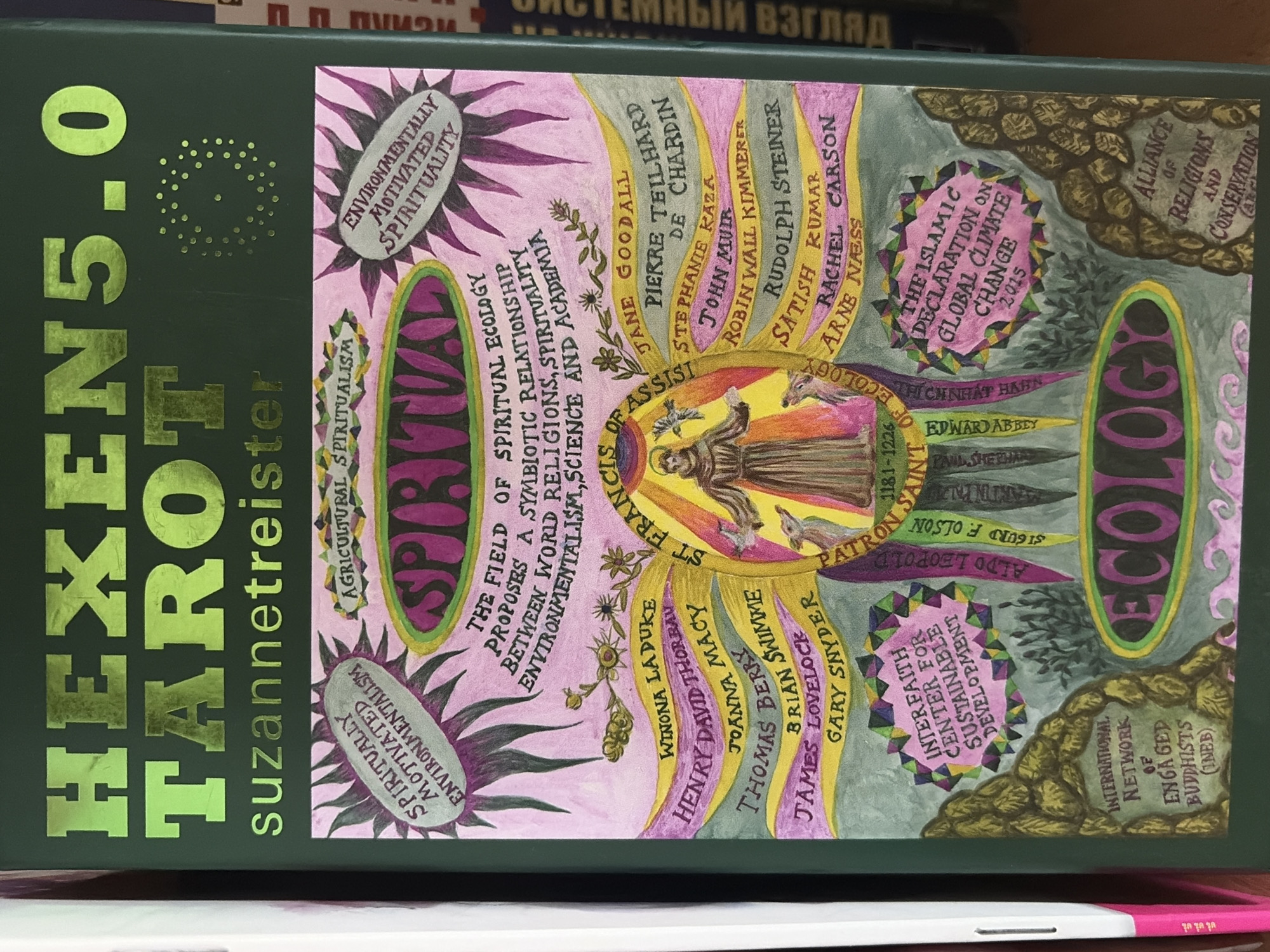 The image displays a close-up of a dark green book or box cover, likely for a tarot deck, titled "HEXENS.0 TAROT" by "suzannetreister," rendered in reflective gold foil. The cover is adorned with a vibrant, hand-drawn illustration centered on the concept of "Spiritual Ecology."
At the heart of the illustration is a figure resembling St. Francis of Assisi, identified as "PATRON SAINT 1181-1226," surrounded by birds and animals. The artwork features sunbursts, leaf-like banners, and organic motifs in shades of pink, purple, yellow, green, and brown.
The text elaborates on Spiritual Ecology as a "symbiotic relationship between world religions, spirituality, environmentalism, science and academia," encompassing themes like "Environmentally Motivated Spirituality" and "Agricultural Spiritualism." Numerous prominent names associated with environmentalism, spirituality, and interfaith efforts are listed, including Jane Goodall, Pierre Teilhard de Chardin, John Muir, Rachel Carson, Arne Ness, Robin Wall Kimmerer, Winona LaDuke, Henry David Thoreau, Joanna Macy, Thomas Berry, James Lovelock, and Gary Snyder, among others.
Also visible are mentions of significant initiatives and organizations such as "The Islamic Declaration on Global Climate Change 2015," "Alliance of Religions and Conservation (ARC)," "Interfaith Center for Sustainable Development," and "International Network of Engaged Buddhists (INEB)." The background is blurred, showing other items, possibly books, on a shelf, with some Russian text ("МНЫЙ ВЗГЛЯД") discernable on one.