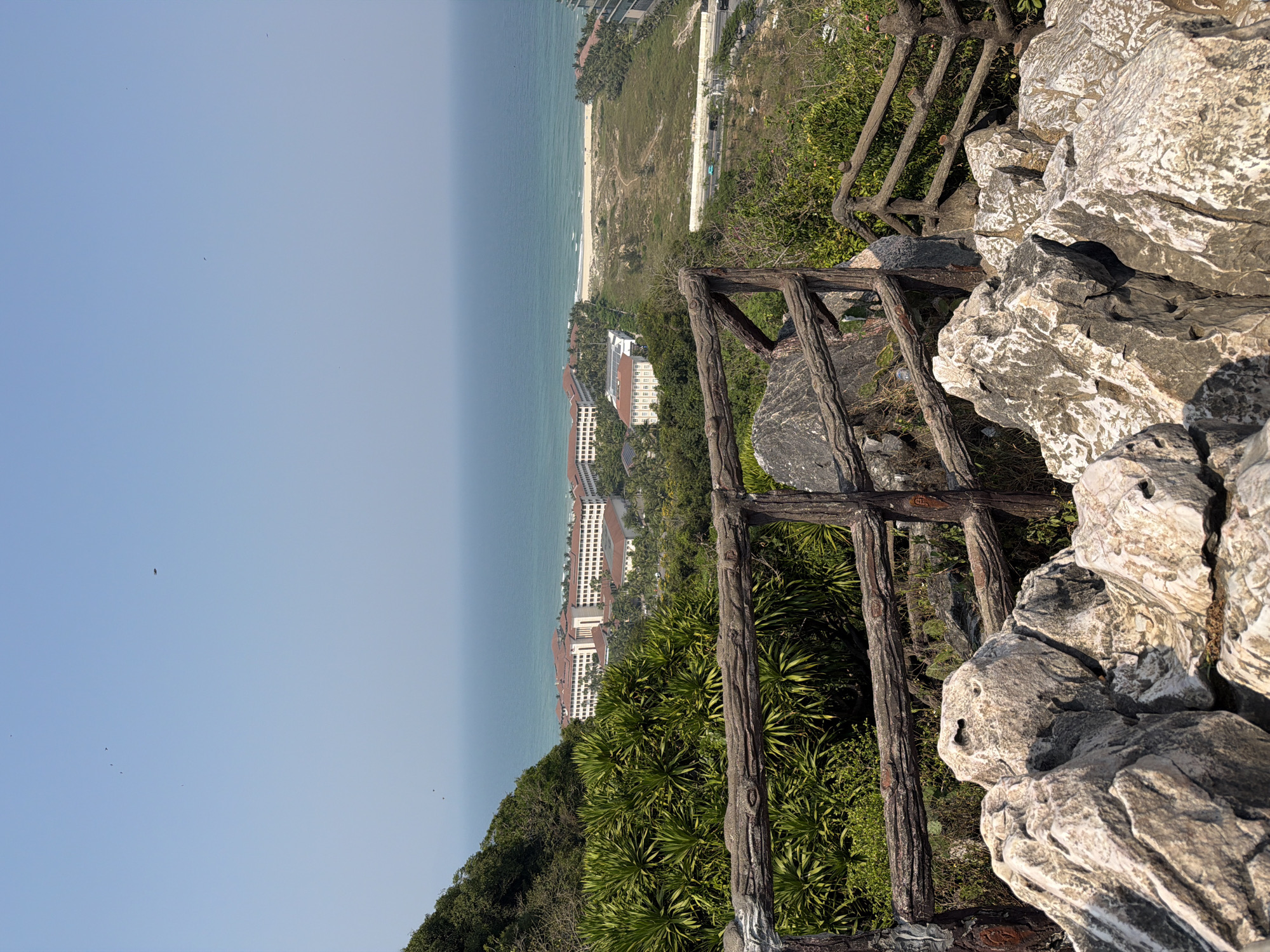 The scene is a high-angle view from a mountainous terrain, likely a viewpoint in Đà Nẵng, Vietnam. The sky is light blue. The ocean meets the shore, where a sandy beach is visible. Buildings are situated near the coastline. A rustic wooden fence borders the cliff edge. Rocks are prominent in the foreground. Green vegetation covers the hillside.