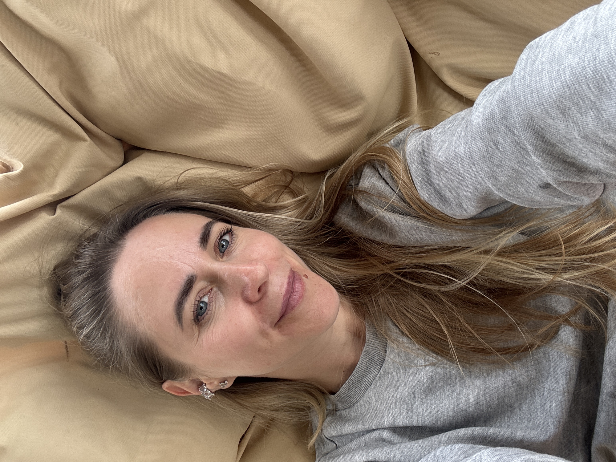 A woman with blonde hair, blue eyes, and a fair complexion lies on a tan-colored surface, likely a bed or couch. She is wearing a grey sweatshirt. Her hair is long and slightly wavy, framing her face. She has on earrings and a ring on her left hand. The woman is looking up towards the camera, with a slight smile on her face. A small mole is visible on her cheek. The lighting suggests it could be daytime, with soft, diffused light. The background is out of focus and appears to be made of fabric, possibly the bedding or upholstery of the furniture she is on. The overall mood of the image is relaxed and casual, as if she is taking a selfie from a comfortable position.