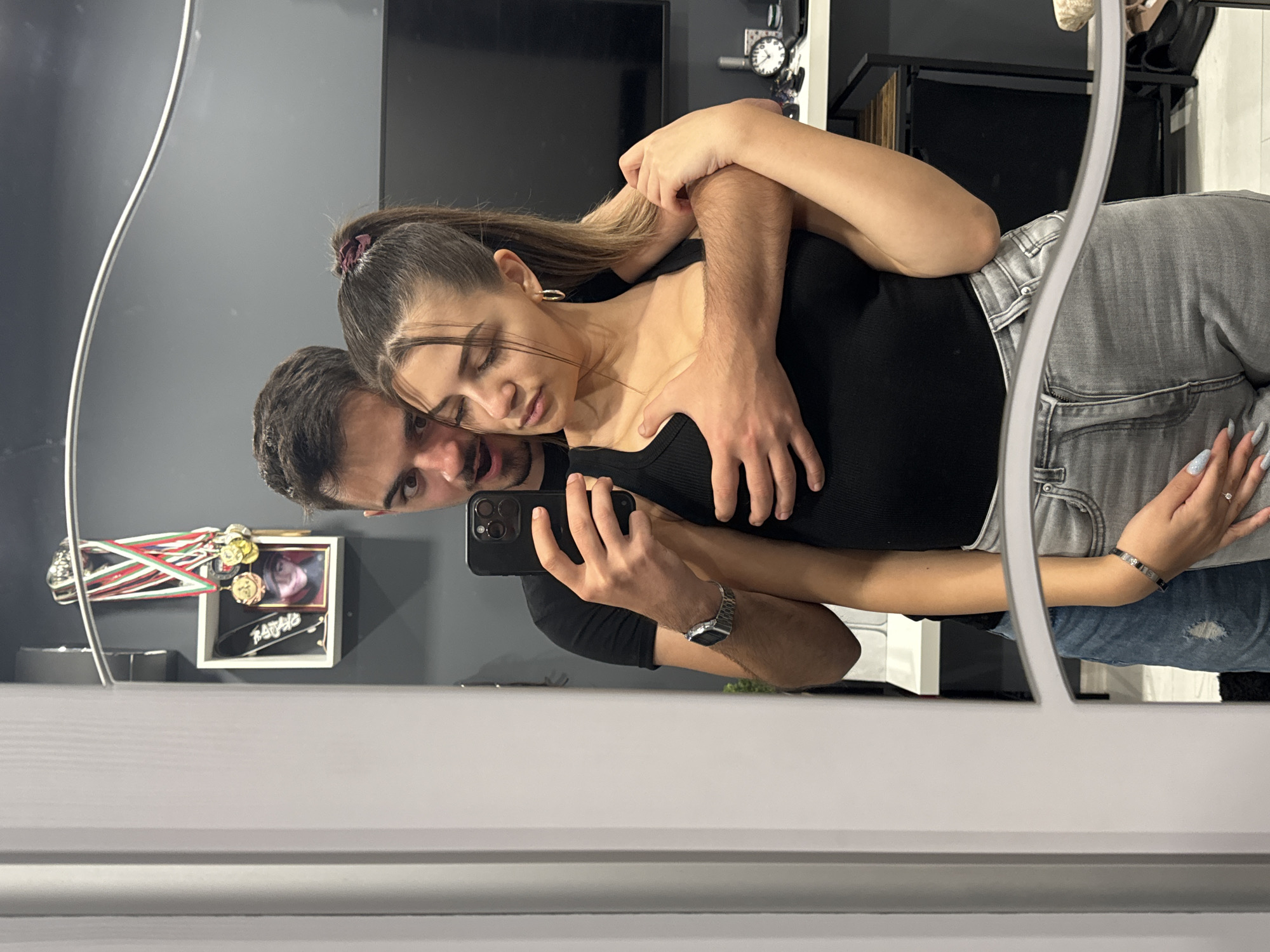 A mirror selfie depicts a man embracing a woman. The woman wears a black top and grey jeans. The man, behind her, holds a black smartphone, taking the picture. His arms are wrapped around her, and he wears a black shirt and a silver watch. They are indoors, possibly in a house or apartment. In the background is a grey wall, a television, and some decorative items, including what appears to be a photo frame and medals. I have no information available about the location being in the city of Sofia, Bulgaria.
