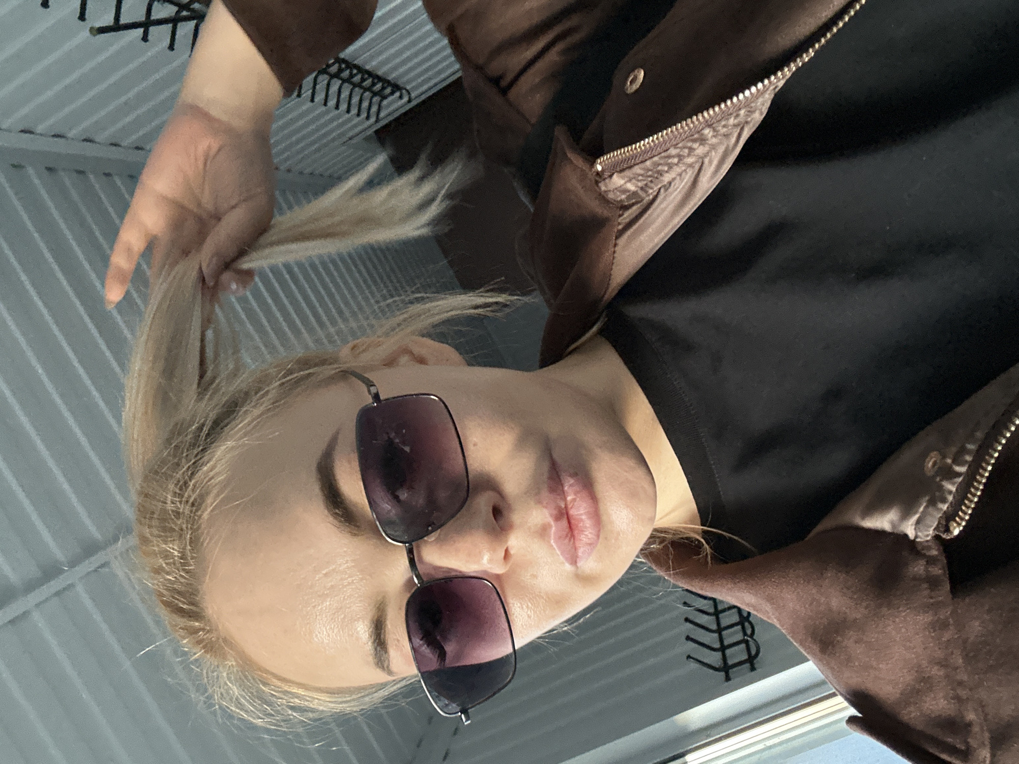 A person with light-blonde hair is positioned at an oblique angle, facing the camera, with their head tilted to the left. They are wearing sunglasses with dark lenses. Their left hand is raised, holding a section of their hair. They are wearing a dark shirt and a brown jacket. The background suggests an indoor setting, possibly with a gray metal grid structure visible overhead. The photo was taken in the city of Megion, Russia.