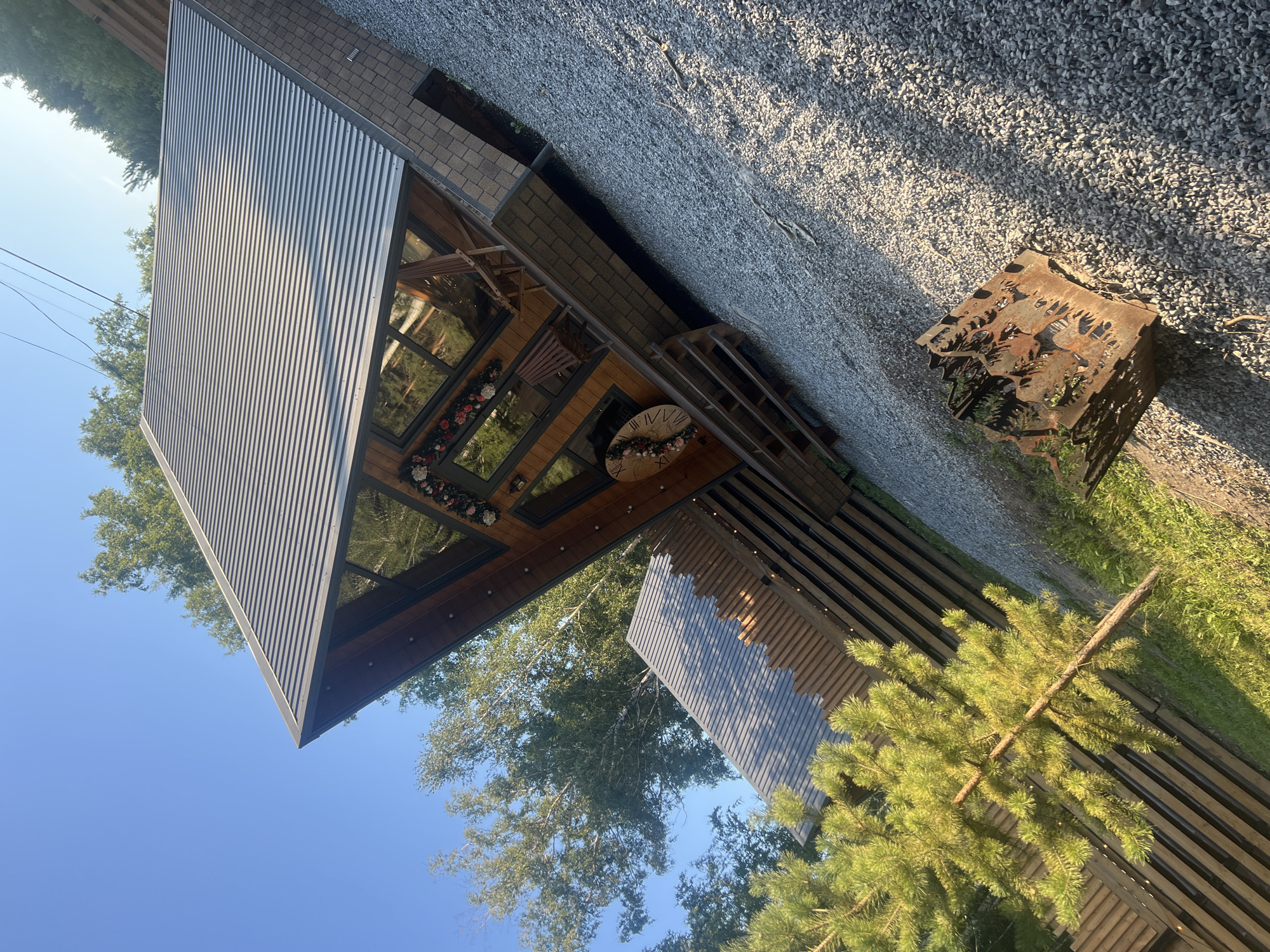 A triangular, modern cabin with a corrugated metal roof and wood siding is situated in a wooded area. The cabin features large windows that reflect the surrounding trees and a bright blue sky. A wooden deck with chairs extends from the front of the cabin. A rustic, metal fire pit is located on a gravel path in the foreground. The scene is outdoors, with lush green trees and grass visible. The image appears to be taken in Omsk, Russia, based on the presence of birch trees commonly found in the region.