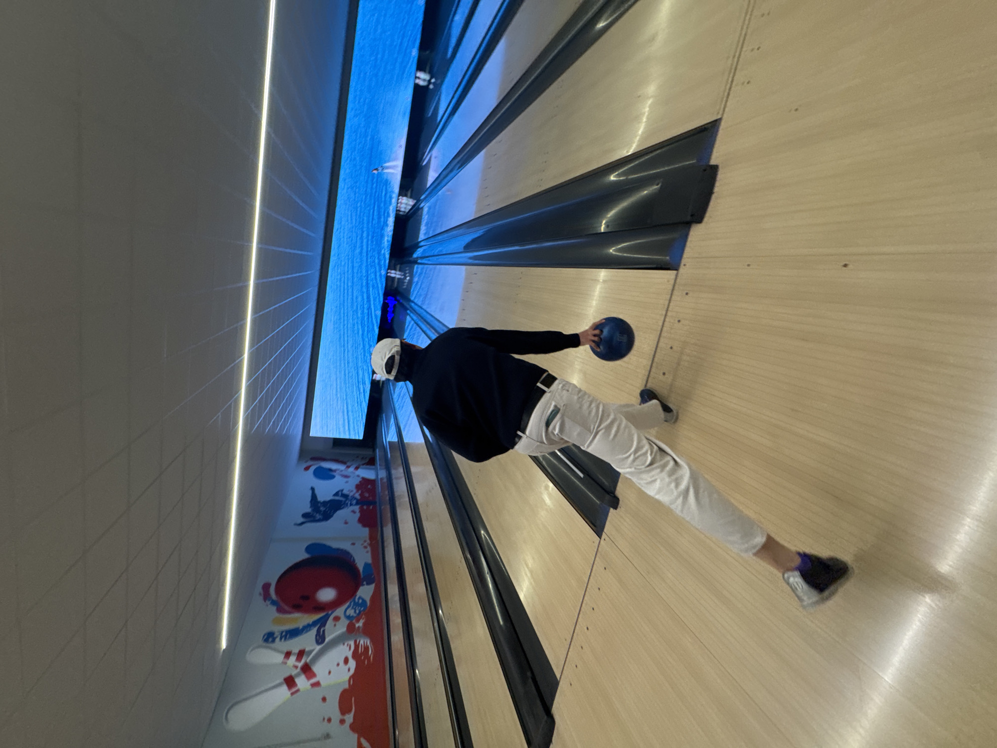 A person is bowling. The person wears a dark shirt, white pants, a white cap, and bowling shoes, and holds a blue bowling ball. The scene takes place in a bowling alley. A monitor displaying an ocean scene is visible along the wall. A painted mural is also visible on the wall.