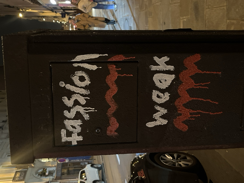 The image captures a dark-colored utility box positioned on a paved sidewalk in Paris, France, at night. The box is defaced with white and red graffiti. In white paint, the word "Fassion" is written horizontally near the top, exhibiting dripping effects. Below it, a wavy horizontal line in red paint also shows drips. Further down, the word "weak" is written in white paint with similar dripping, followed by another wavy red line with drips at the bottom of the box.
To the left of the utility box, a street is visible with two parked vehicles: a white SUV partially obscured by the box, and a dark-colored sedan, of which only the front-left portion and wheel are visible in the foreground. Behind the street and vehicles, multi-story buildings with typical Parisian architectural elements, including balconies and arched windows, line the thoroughfare. One building displays a sign that reads "VIRTUO."
To the right of the utility box, a stone-paved sidewalk recedes into the background. Two individuals are seen walking away from the viewer on this sidewalk. The person on the left wears a long, light brown trench coat, while the person on the right is dressed in a light-colored jacket and blue jeans. The background on the right also features additional buildings with illuminated ground-floor windows, suggesting commercial establishments. The overall scene is illuminated by artificial streetlights under a dark night sky.