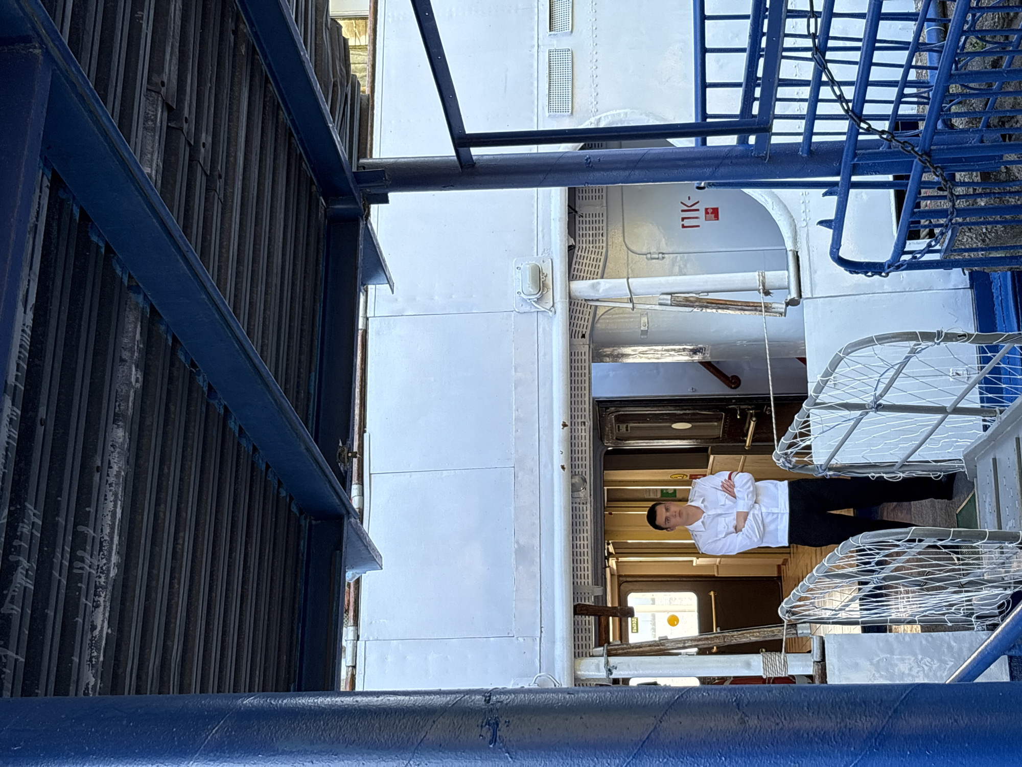 A young man stands with his arms crossed, facing forward, on a pier or dock in Chaikovsky, Russia. He is wearing a white collared shirt with red armbands and dark pants. The man is positioned on a staircase leading towards an entrance of a large, white vessel, possibly a ferry or boat. The vessel has a dark, slatted roof overhead, supported by blue metal beams. White metal railings with netting line the staircase. The background reveals an open doorway into the vessel with a sign that reads "ВЫХОД" (EXIT) in Cyrillic. The overall environment suggests an outdoor maritime setting.