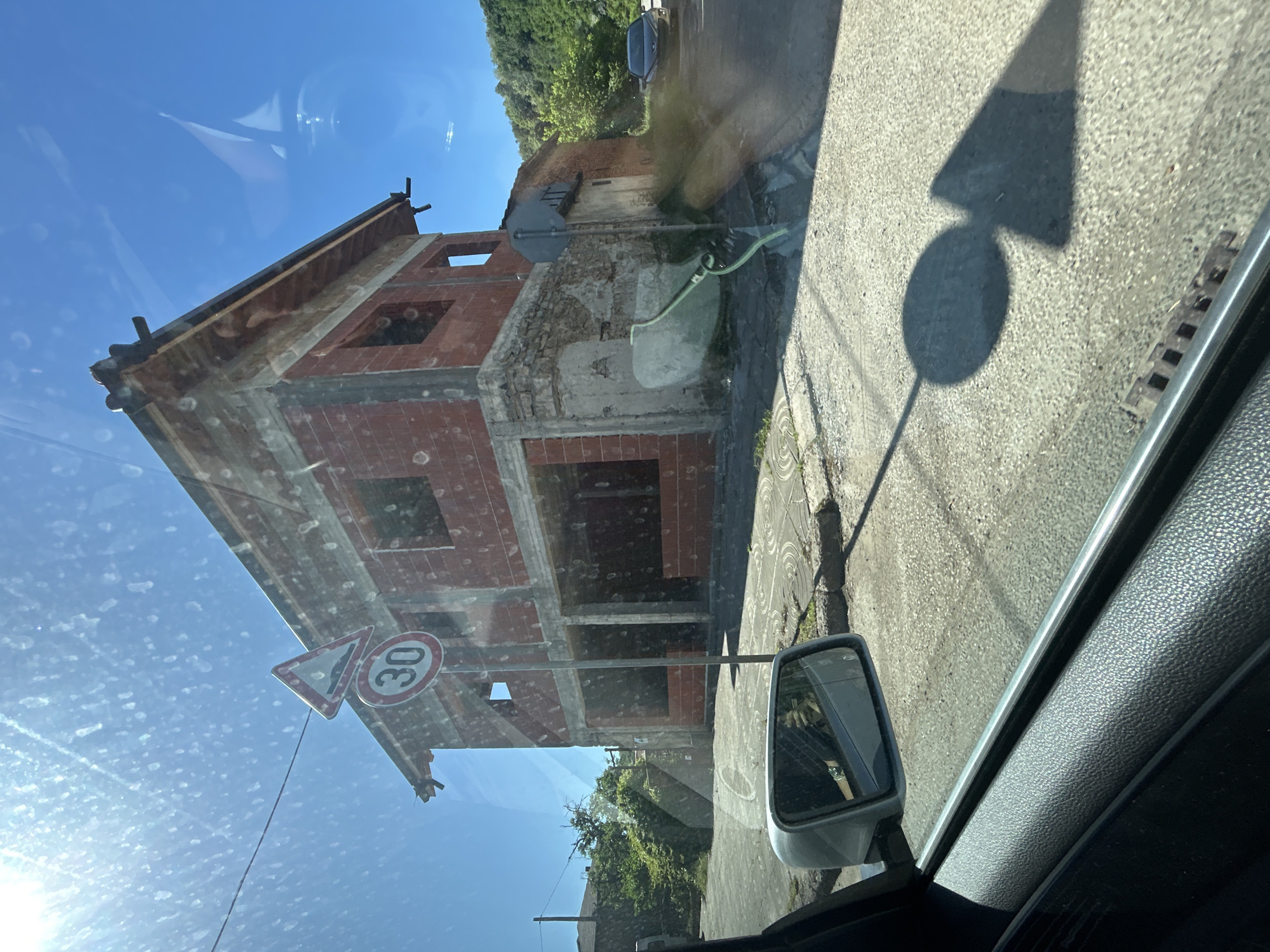 The image appears to be taken from inside a vehicle, possibly a car. The view is of an unfinished or dilapidated brick building located in Balgarski Izvor, Bulgaria, as a street sign near the building indicates a speed limit of 30 (likely km/h). The structure is a two-story building with no windows or doors, and a partially ruined roof. A shadow of a street sign is cast on the road. A side mirror is visible, reflecting the environment. The car’s dashboard is visible in the foreground. The sky is visible through the car's window, and there are water spots on the window glass.