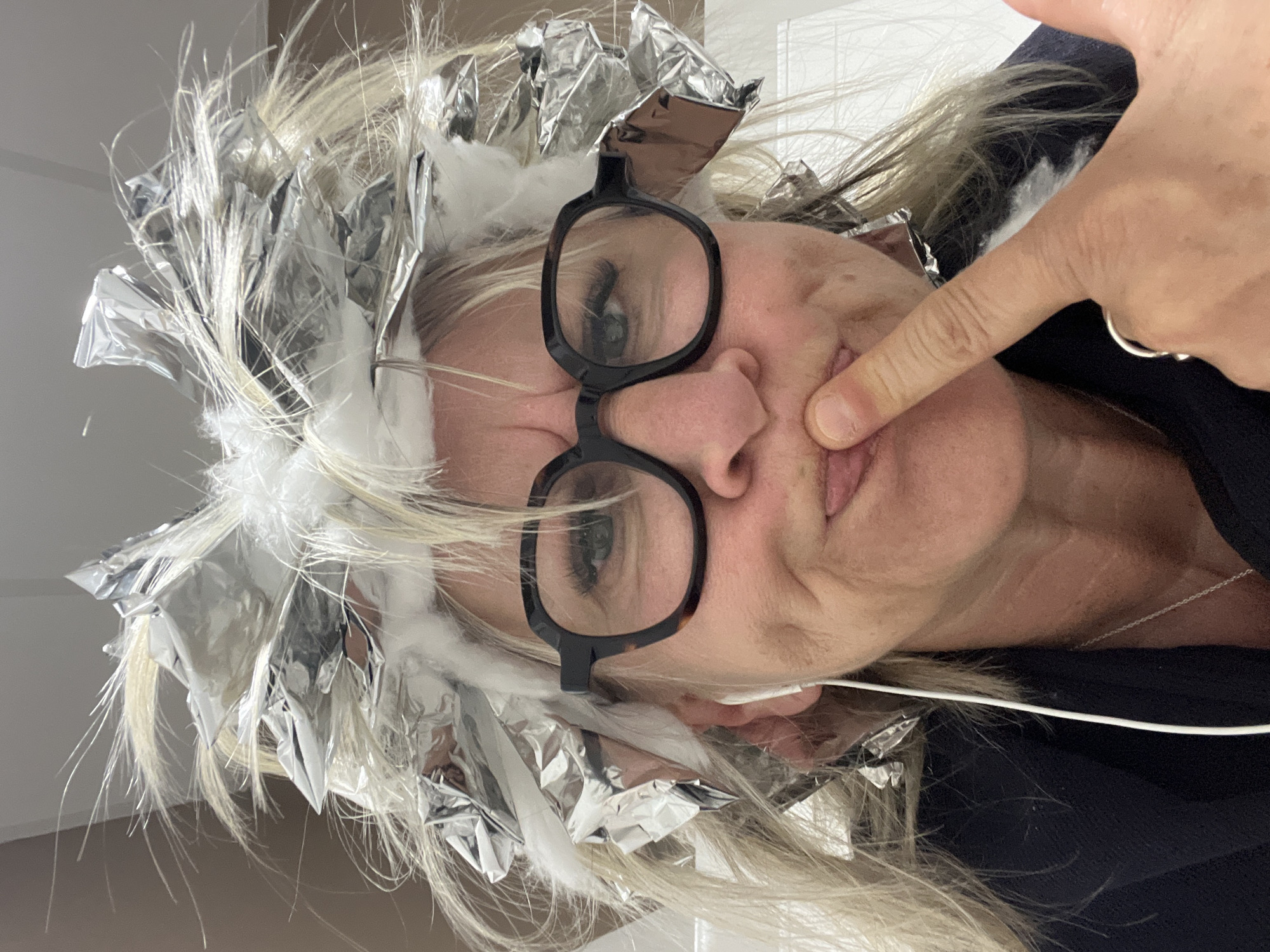 A middle-aged person wearing glasses and with aluminum foil and cotton strips in their blonde hair is shown in a close-up selfie-style photograph. The person's index finger, adorned with a ring, touches their cheek. A white earbud cord is visible near their mouth. The person wears a dark-colored top. No specific location is identifiable in the image, and there is no discernible connection to Montrond-les-Bains, France.