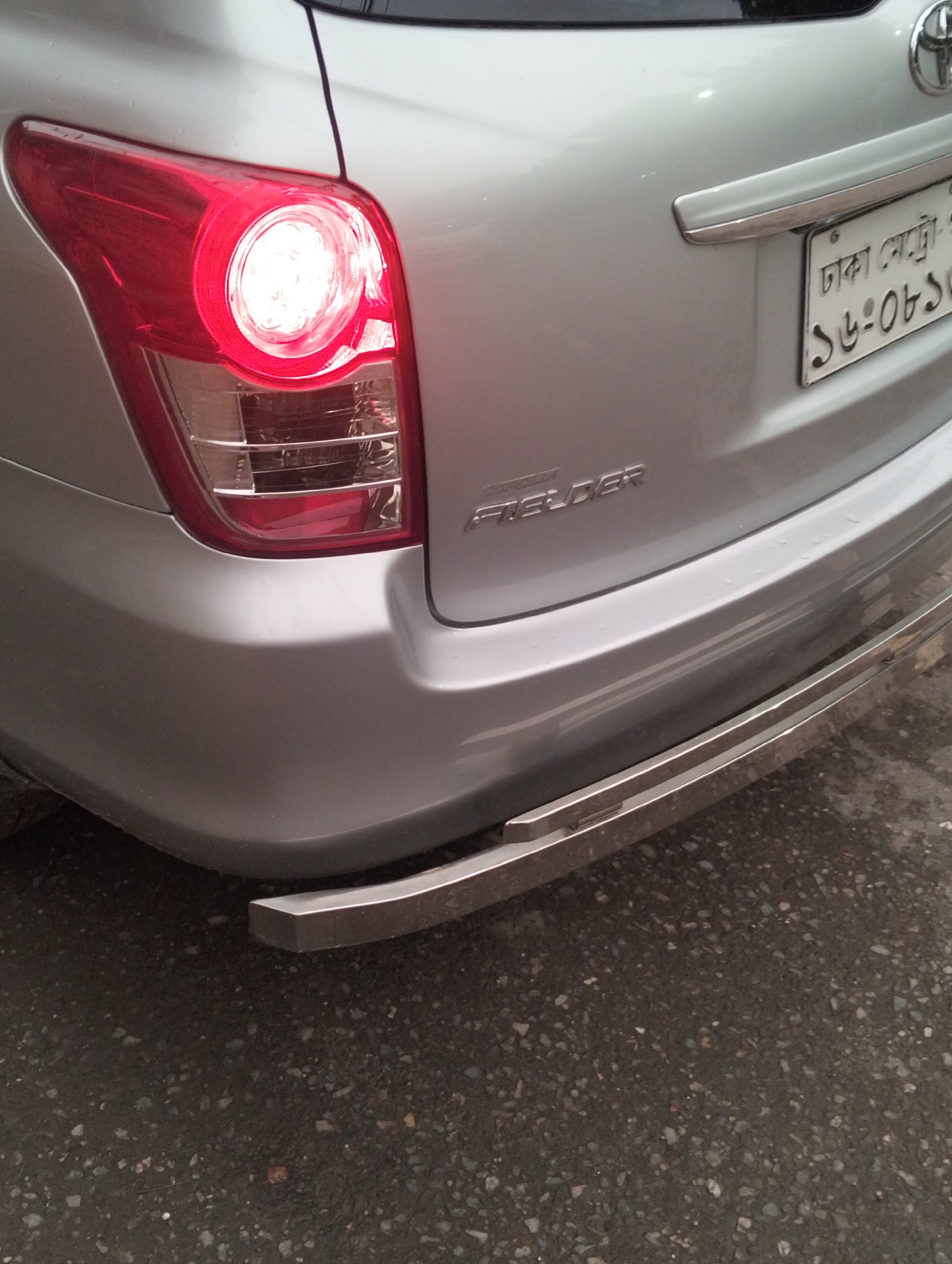 The image captures the rear left section of a silver Toyota Corolla Fielder, parked on an asphalt surface. The car's left taillight is prominently illuminated, showcasing a circular cluster of bright lights within a red housing, suggesting either the brake light is engaged or the parking lights are on. Below this, clear segments are visible for other lighting functions.
The silver body of the car appears slightly wet with small water droplets, indicating recent rain or dew. A chrome strip runs across the trunk lid above the license plate, which is clearly visible. The license plate displays text in Bengali script, including "ঢাকা মেট্রো" (Dhaka Metro), confirming the location as Dhaka, Bangladesh. Part of the license number "১৮-০৮১" is also legible.
Below the trunk lid, the car's model name, "COROLLA FIELDER," is embossed on the silver bodywork. A chrome, tubular bumper guard is attached to the lower part of the silver rear bumper, extending outwards from the car's body. The ground beneath the car is a dark, coarse asphalt or tarmac surface. No people or other vehicles are visible, and the scene appears to be set during daytime, possibly overcast, given the lack of harsh shadows.