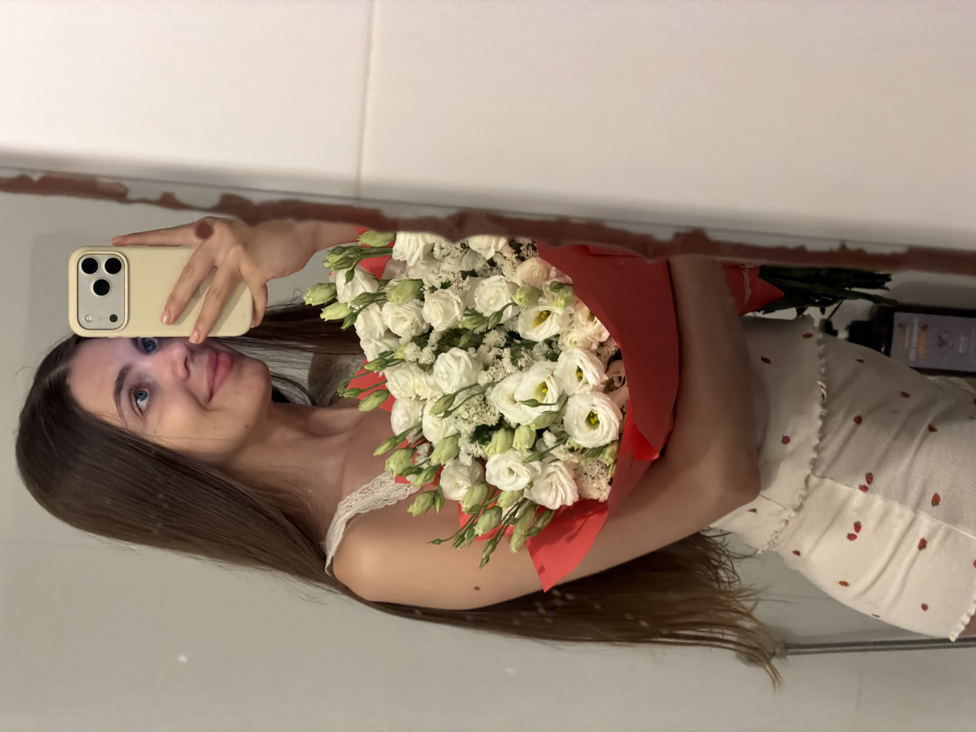 A young woman with long, brown hair is taking a mirror selfie in what appears to be a bathroom. She is holding a bouquet of white roses wrapped in red paper. She is wearing a white top with lace trim and white shorts with a strawberry pattern. The background shows a white tiled wall and a mirror. The location is Rishon LeZion, Israel.